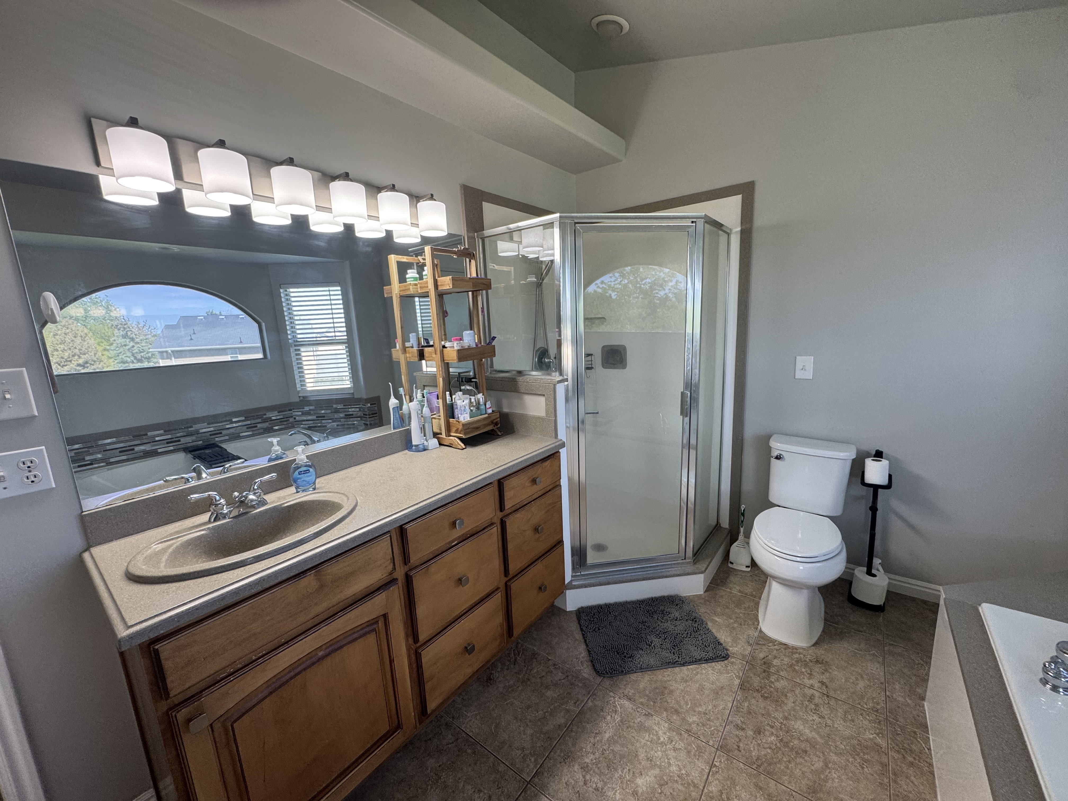 Spacious bathroom featuring a wood-finish vanity with a single basin sink, a large wall-mounted mirror, a multi-light fixture, a corner glass-enclosed shower, and tiled flooring