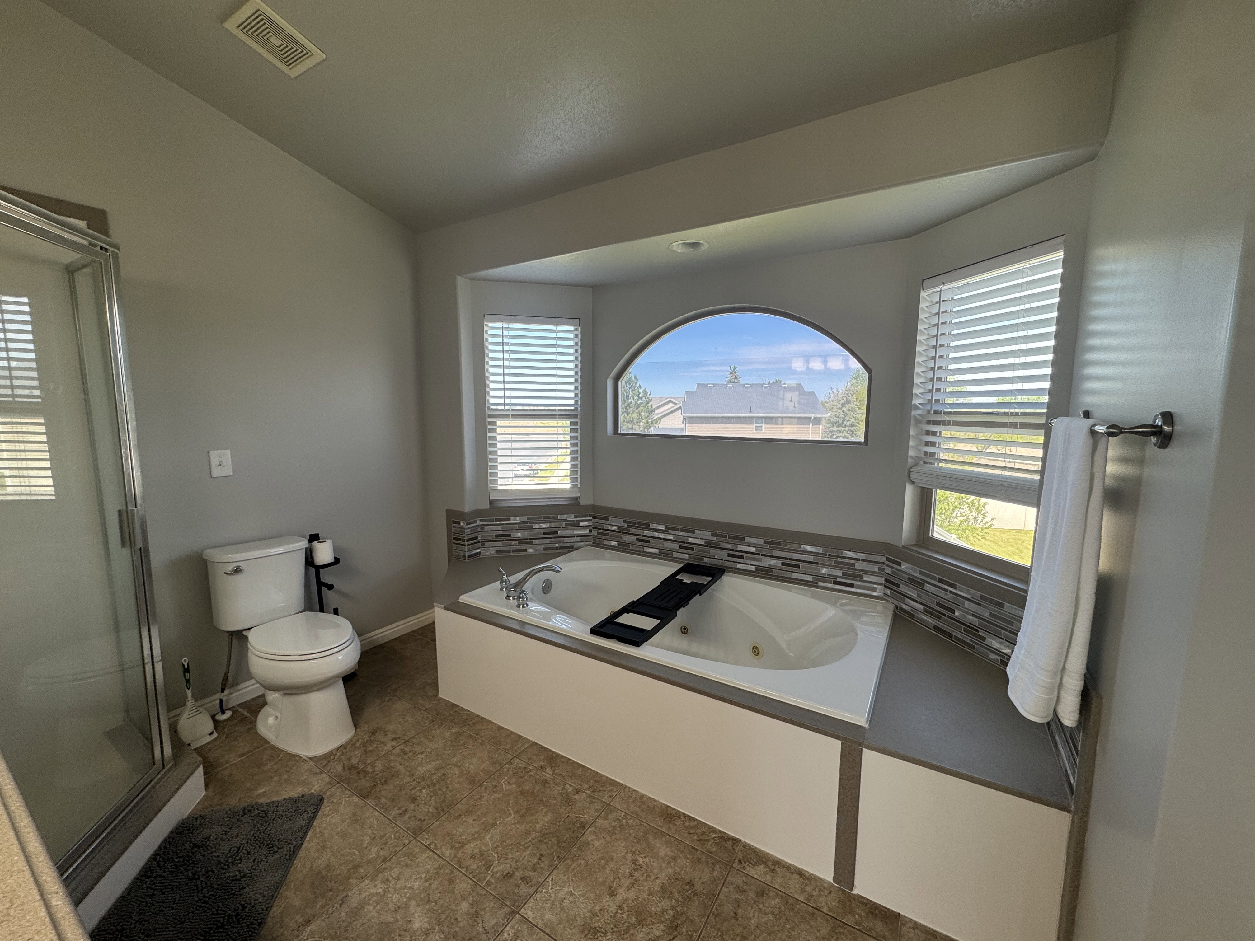 Soaking tub featuring a tiled surround, arched transom window, and adjacent rectangular window with blinds