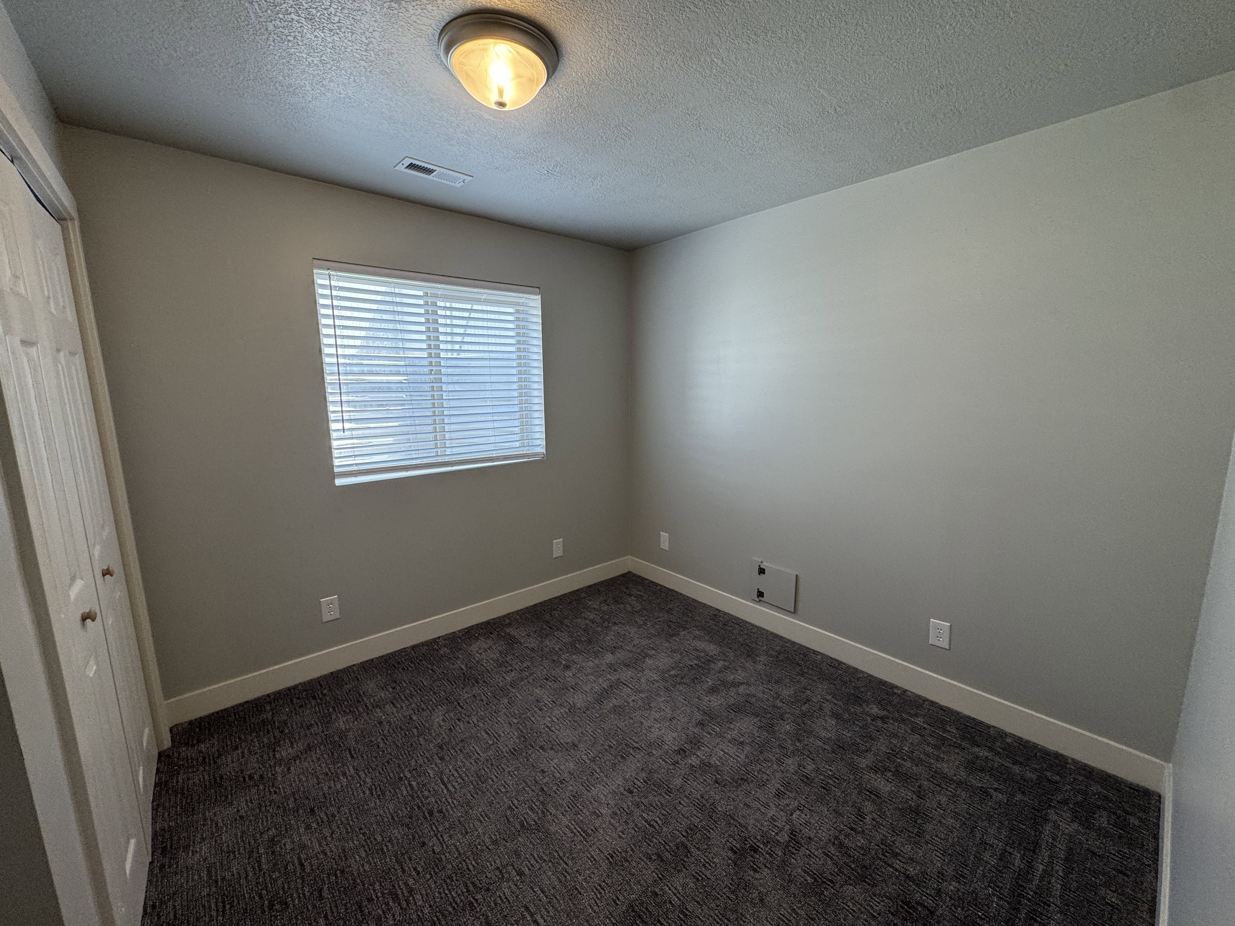 Neutral-toned room featuring a single window with horizontal blinds, overhead flush mount lighting, textured carpeting, and white baseboards