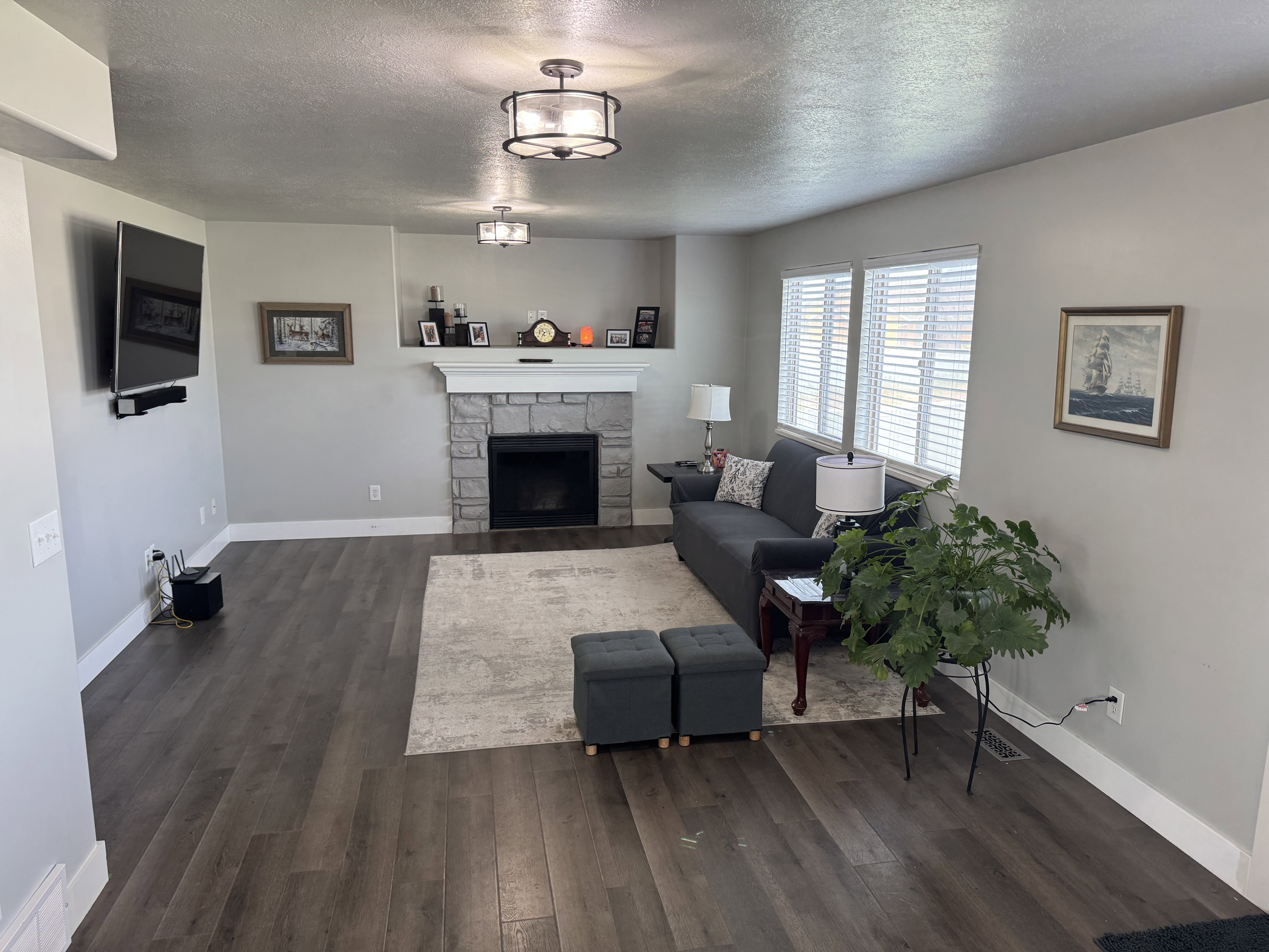 Living area featuring wood-finish flooring, a stone-surround fireplace with a white mantel, and two windows with blinds