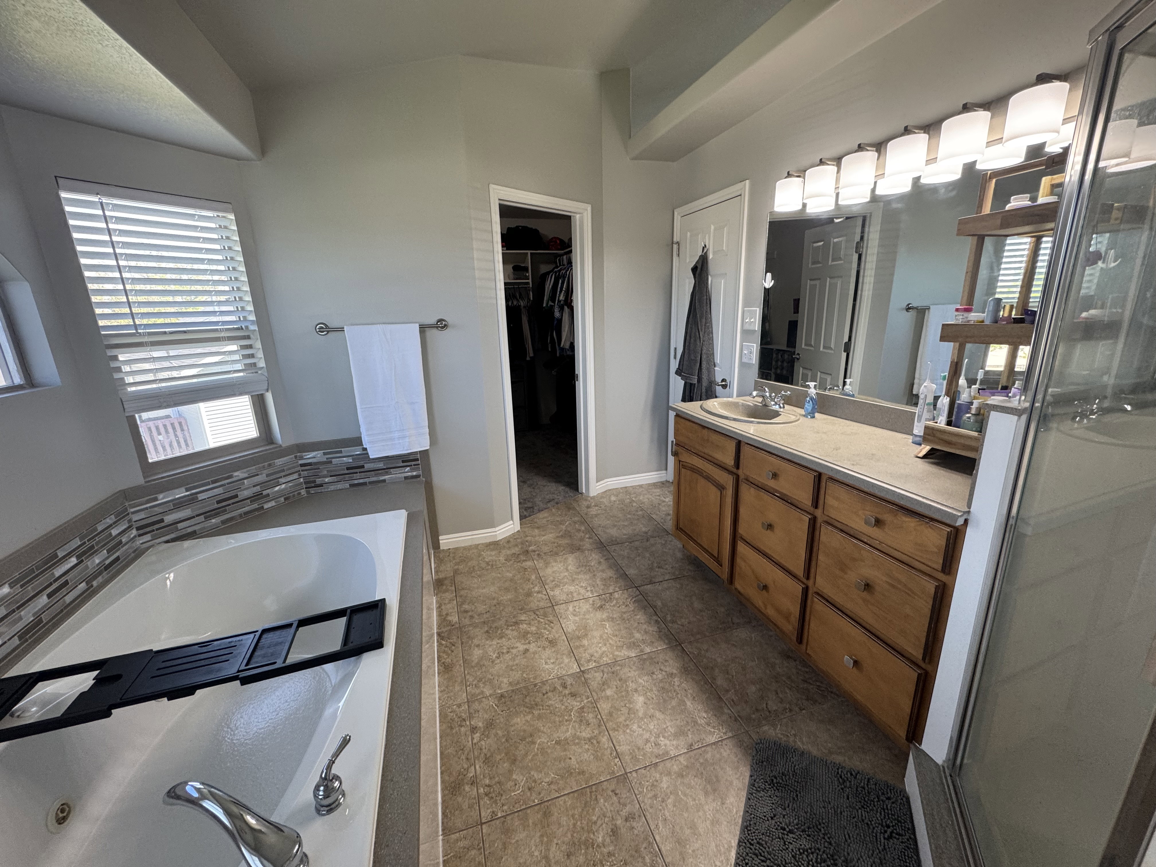 Bathroom featuring a jetted soaking tub with mosaic tile surround, a spacious vanity with wood-finish cabinetry, a large wall-mounted mirror, and tile flooring