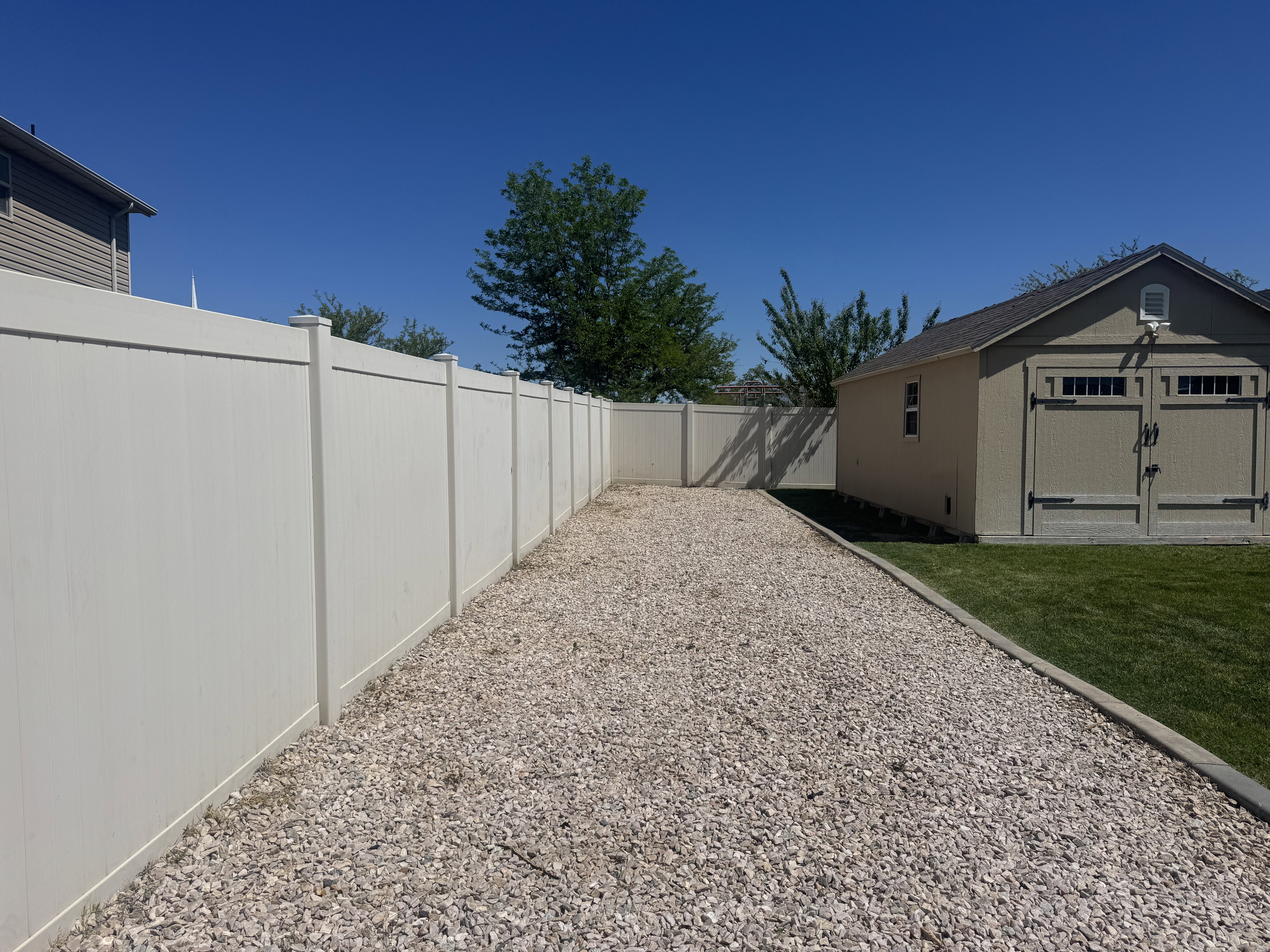 Expansive gravel-covered side yard featuring a tall privacy fence, a detached storage shed, and a manicured lawn border