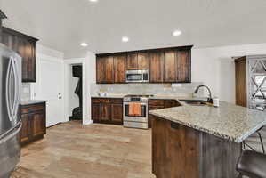 Kitchen featuring a large island with a stone countertop and integrated sink, stainless steel appliances, dark wood cabinetry, a tile backsplash, and recessed lighting