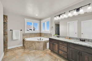 Bathroom featuring a corner soaking tub with natural stone tile surround, dual windows with white plantation shutters, and a vanity with double sinks, light-toned countertop, and dark wood cabinetry