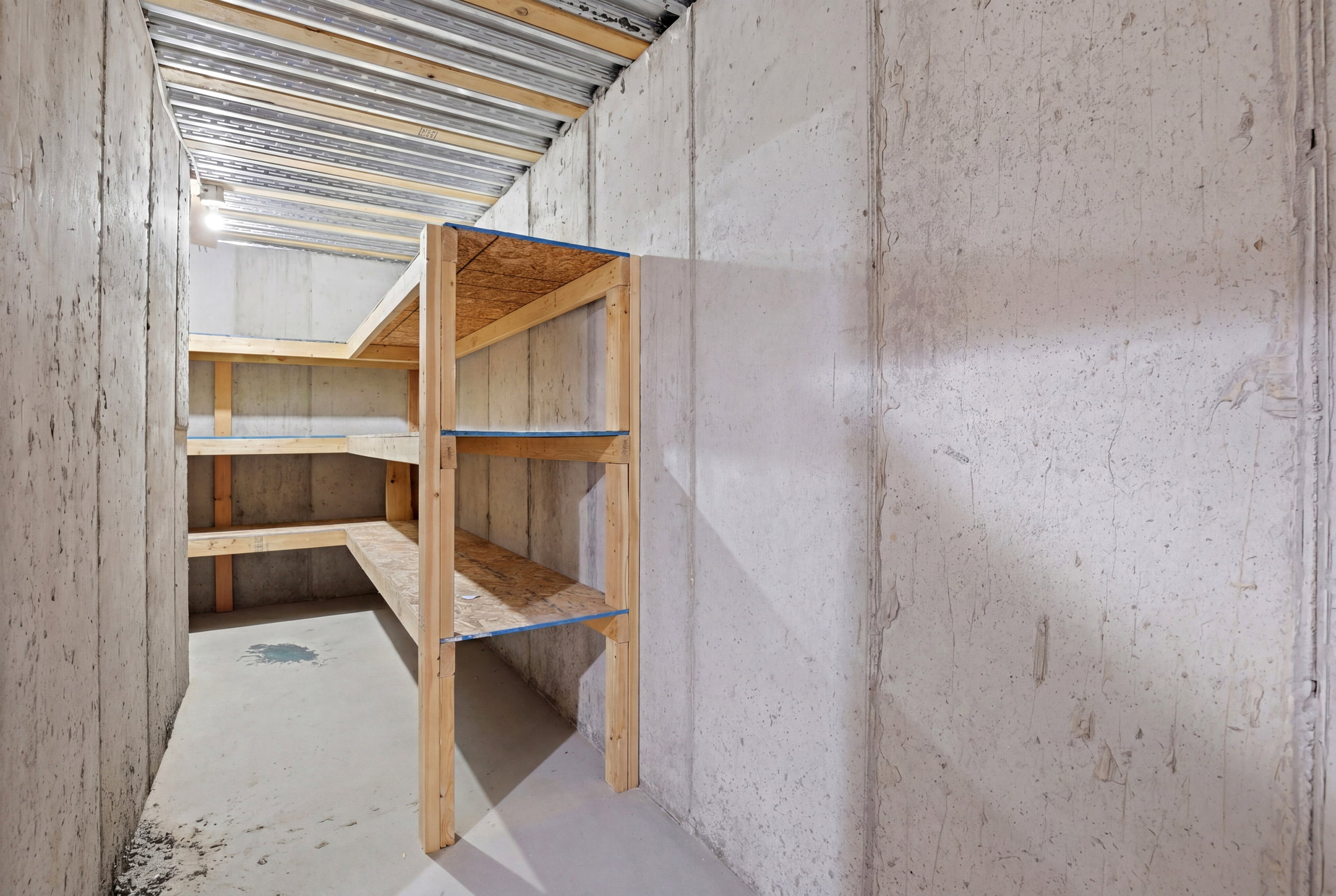 Concrete-walled utility space featuring built-in wooden shelving, a concrete floor, exposed metal ceiling joists, and overhead lighting