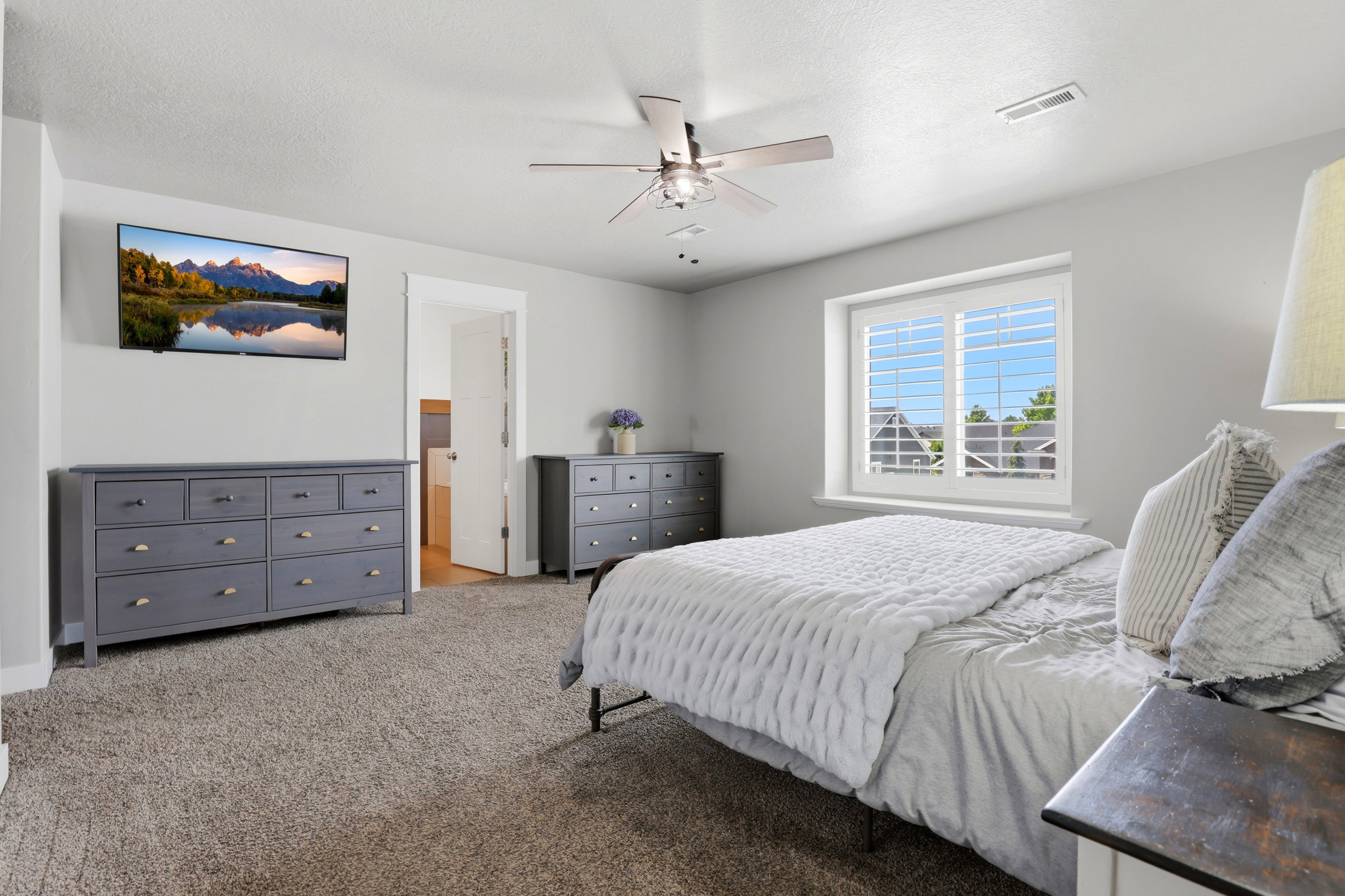 Carpeted room featuring a ceiling fan, a window with white plantation shutters, and a wall-mounted television