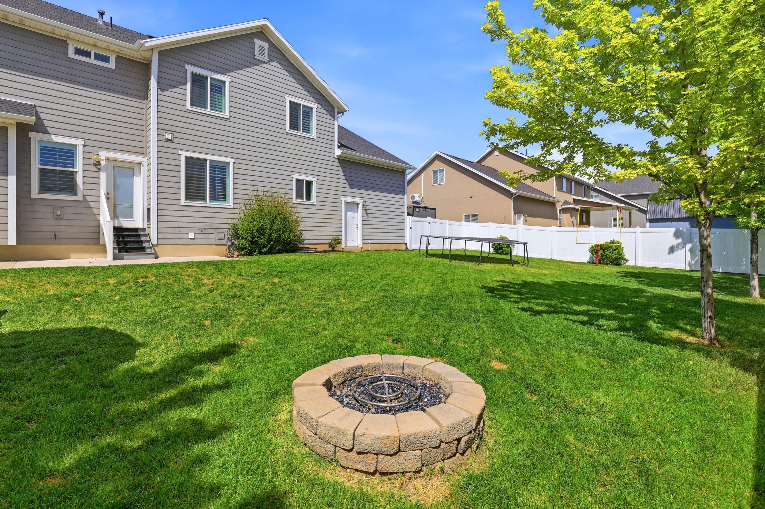 Expansive green lawn featuring a stone-clad fire pit, mature trees, and a white privacy fence