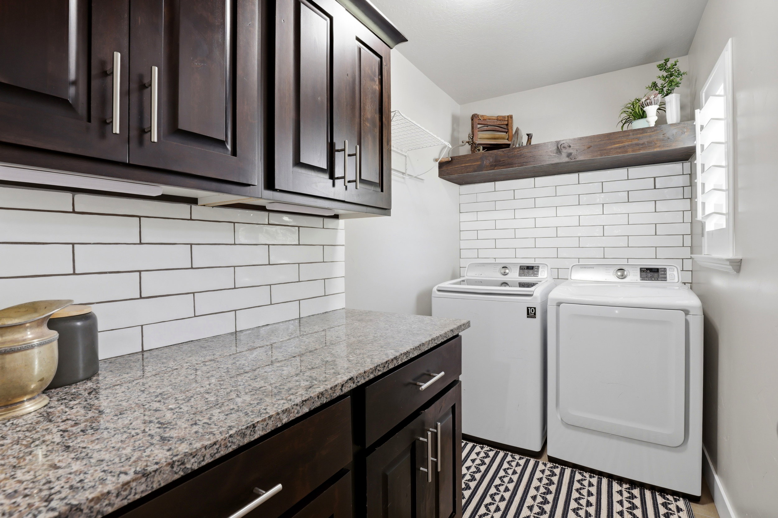 Laundry area featuring dark wood-finish cabinetry, a light granite countertop, white subway tile backsplash, and a window with white shutters