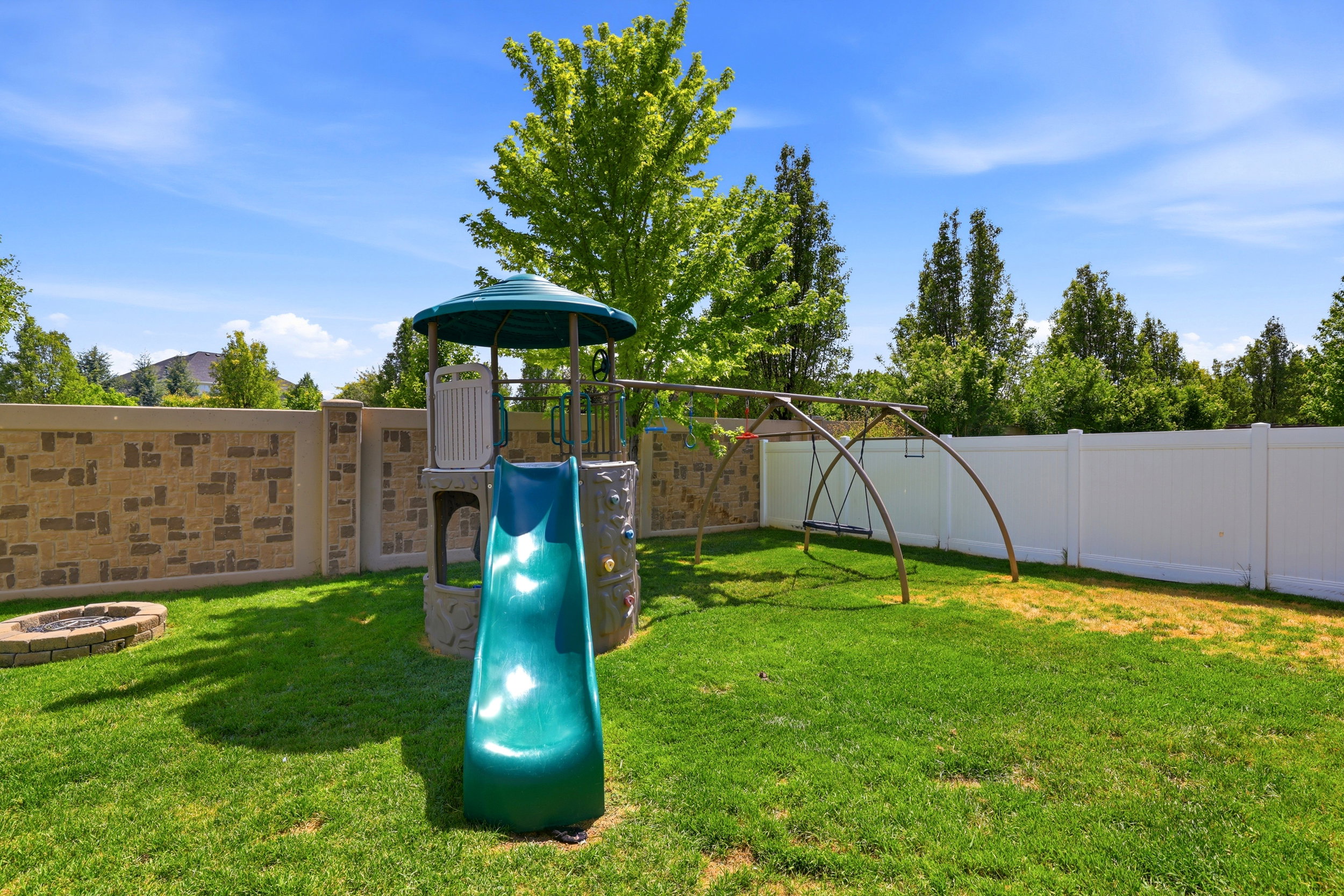 Expansive lawn area featuring a stone-lined fire pit, a stone-patterned privacy wall, and a white vinyl fence