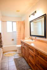 Bathroom featuring a wood-finish vanity with an integrated sink and light-toned countertop, a framed mirror with overhead vanity lighting, a white bathtub with shower, and tiled flooring