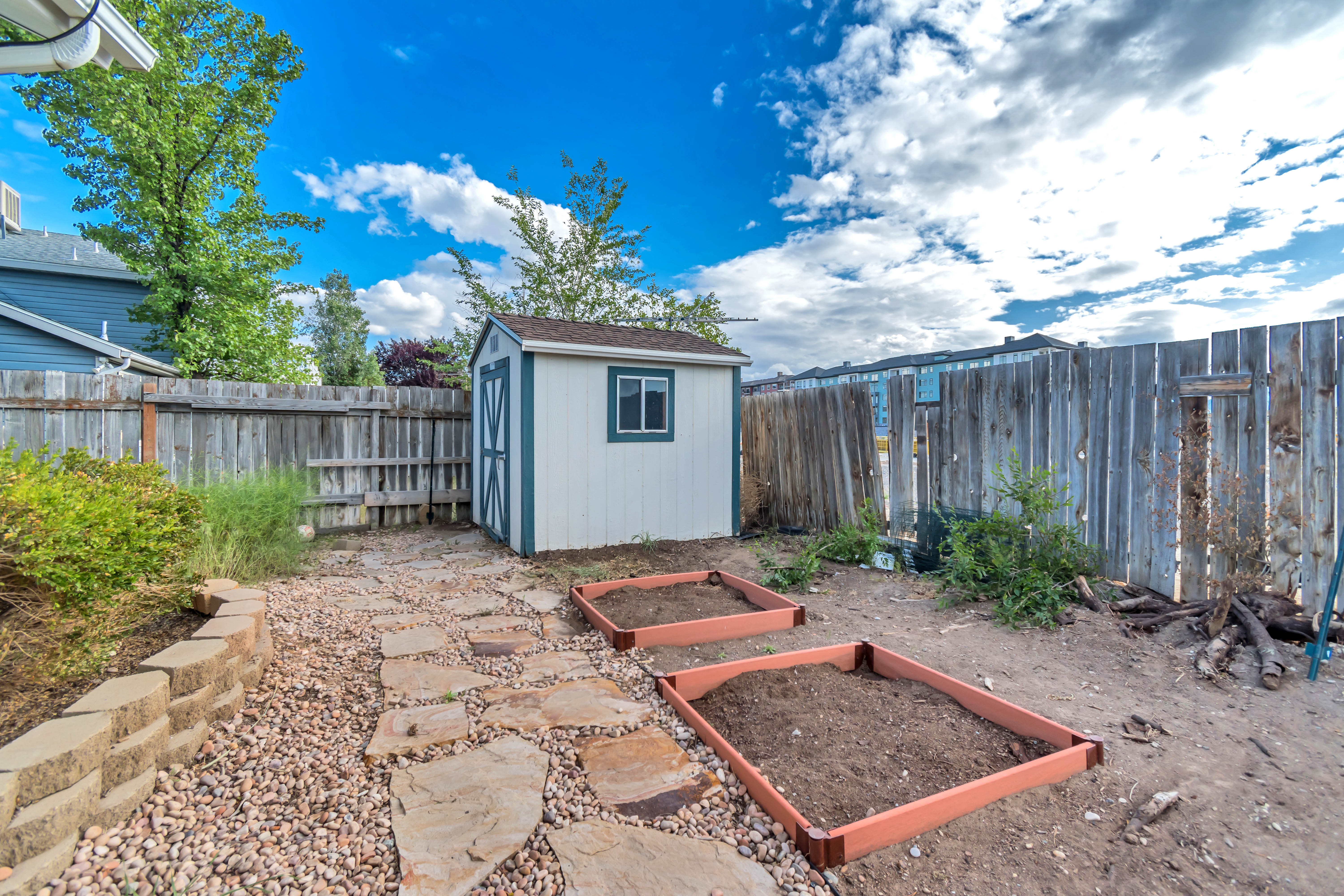 Backyard featuring a white storage shed with teal trim, raised garden beds, a flagstone path, and a retaining wall