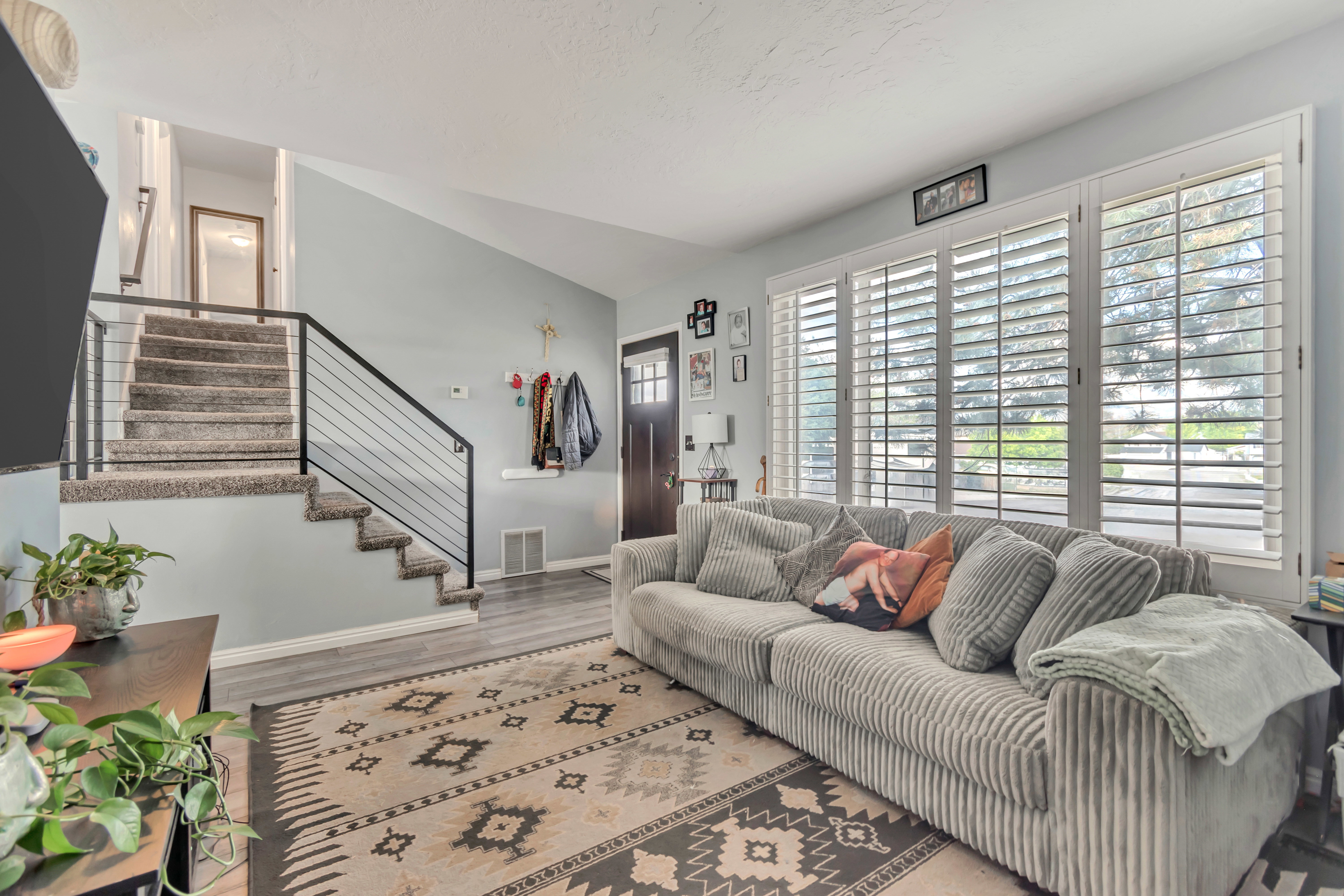 Living area featuring a carpeted staircase with modern black metal railings, wood-finish flooring, a dark entry door, and multiple windows with white plantation shutters