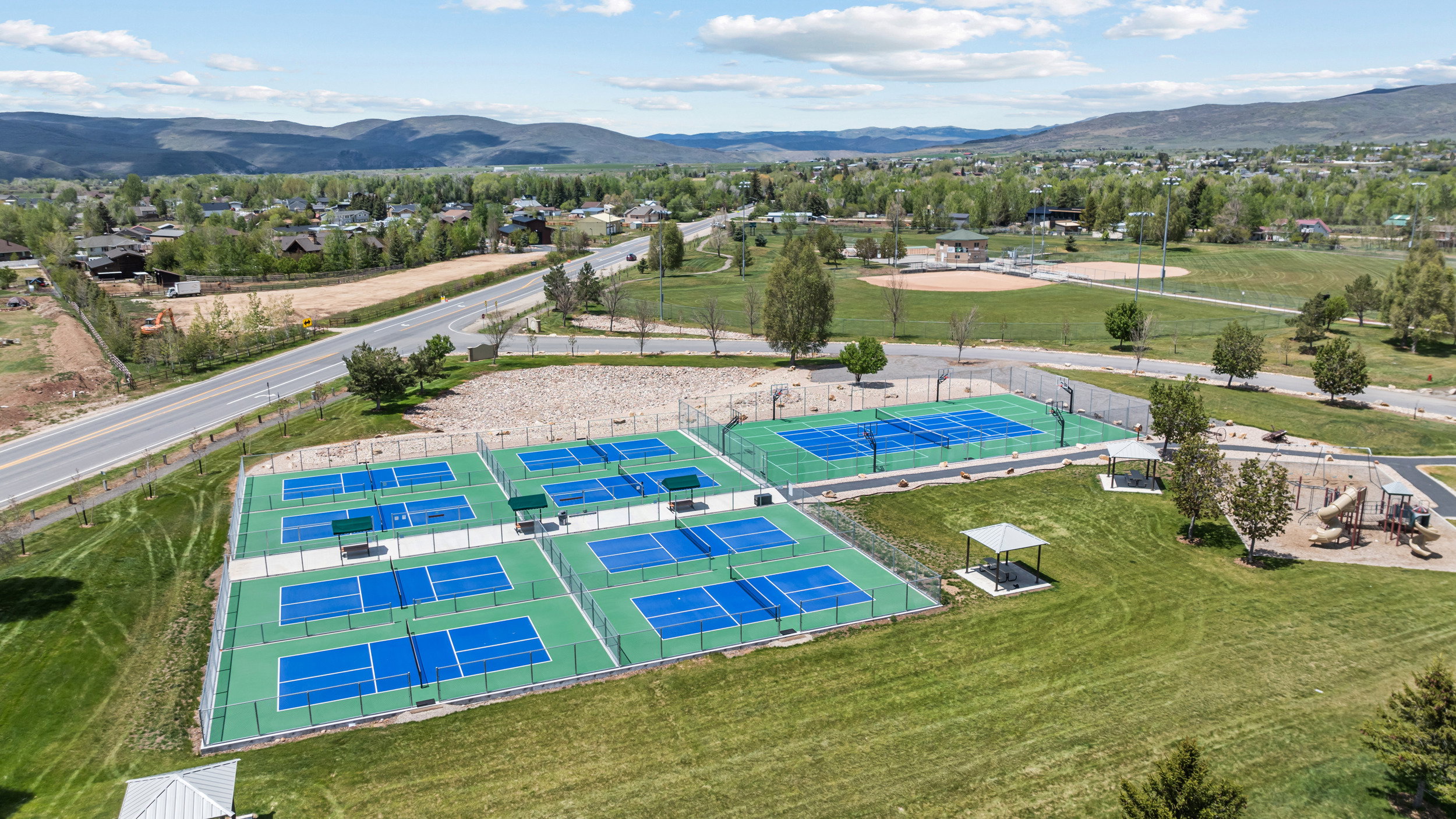 Expansive recreational area featuring multiple blue and green pickleball courts, tennis courts, a baseball field, and a dedicated playground area