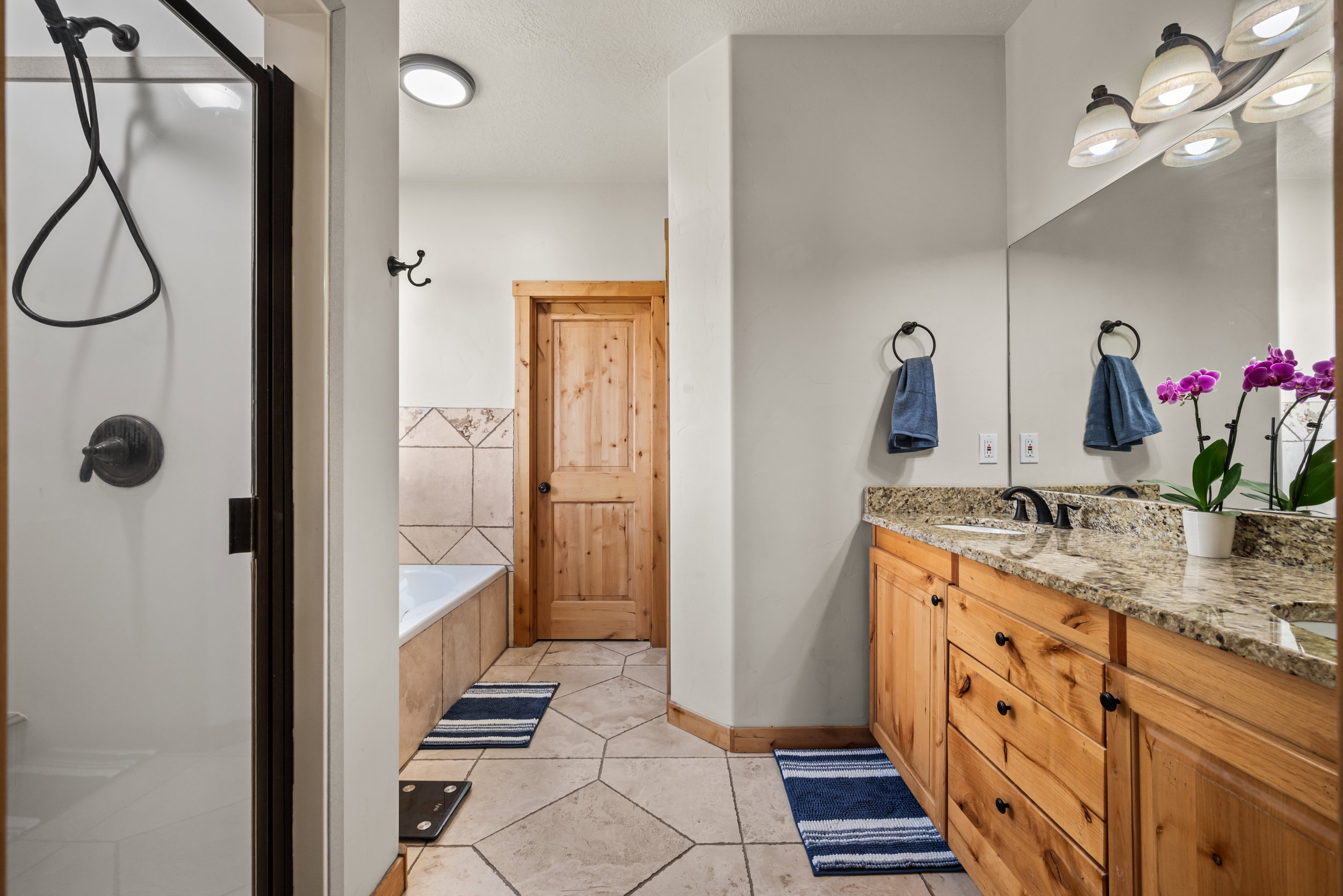 Bathroom featuring a wood-finish vanity with a light-colored countertop, a built-in shower with dark bronze hardware, a built-in bathtub with a tiled surround, a full-width mirror, and large format floor tiles