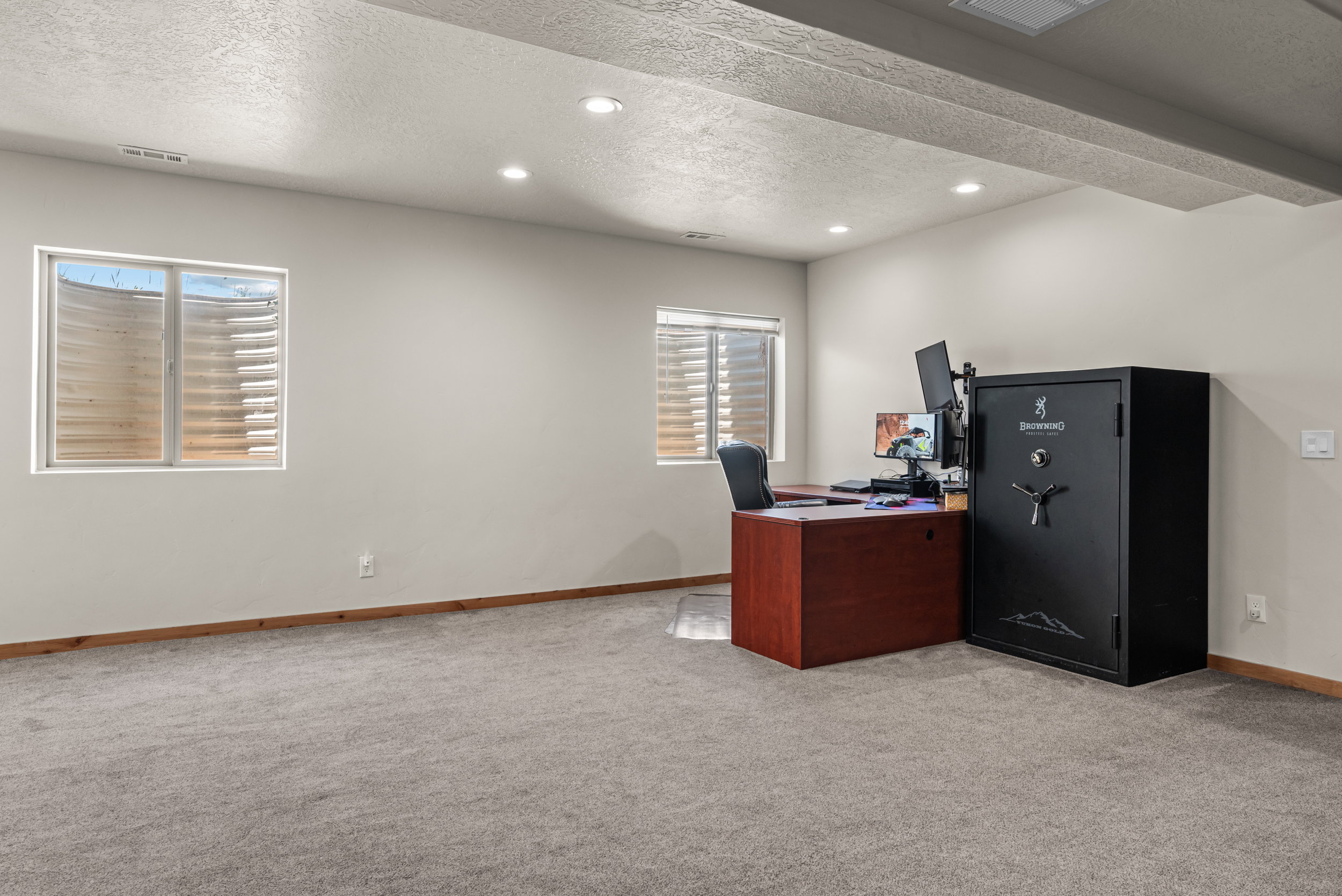 Spacious room featuring light gray carpeting, neutral wall finishes, two egress windows with blinds, wood-finish baseboards, and recessed ceiling lighting