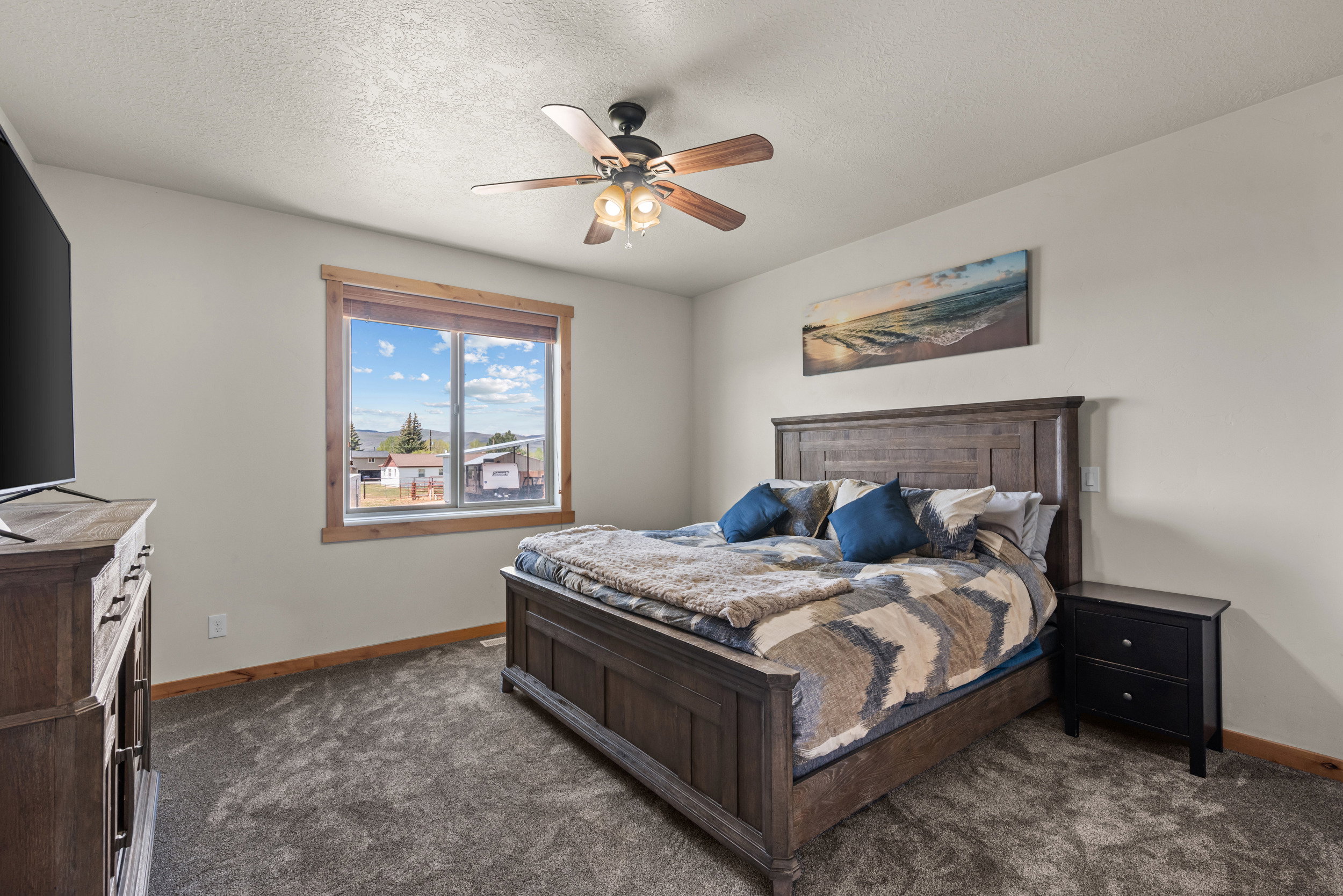 Carpeted bedroom featuring a large window with natural wood trim, a ceiling fan with light fixture, and neutral wall paint