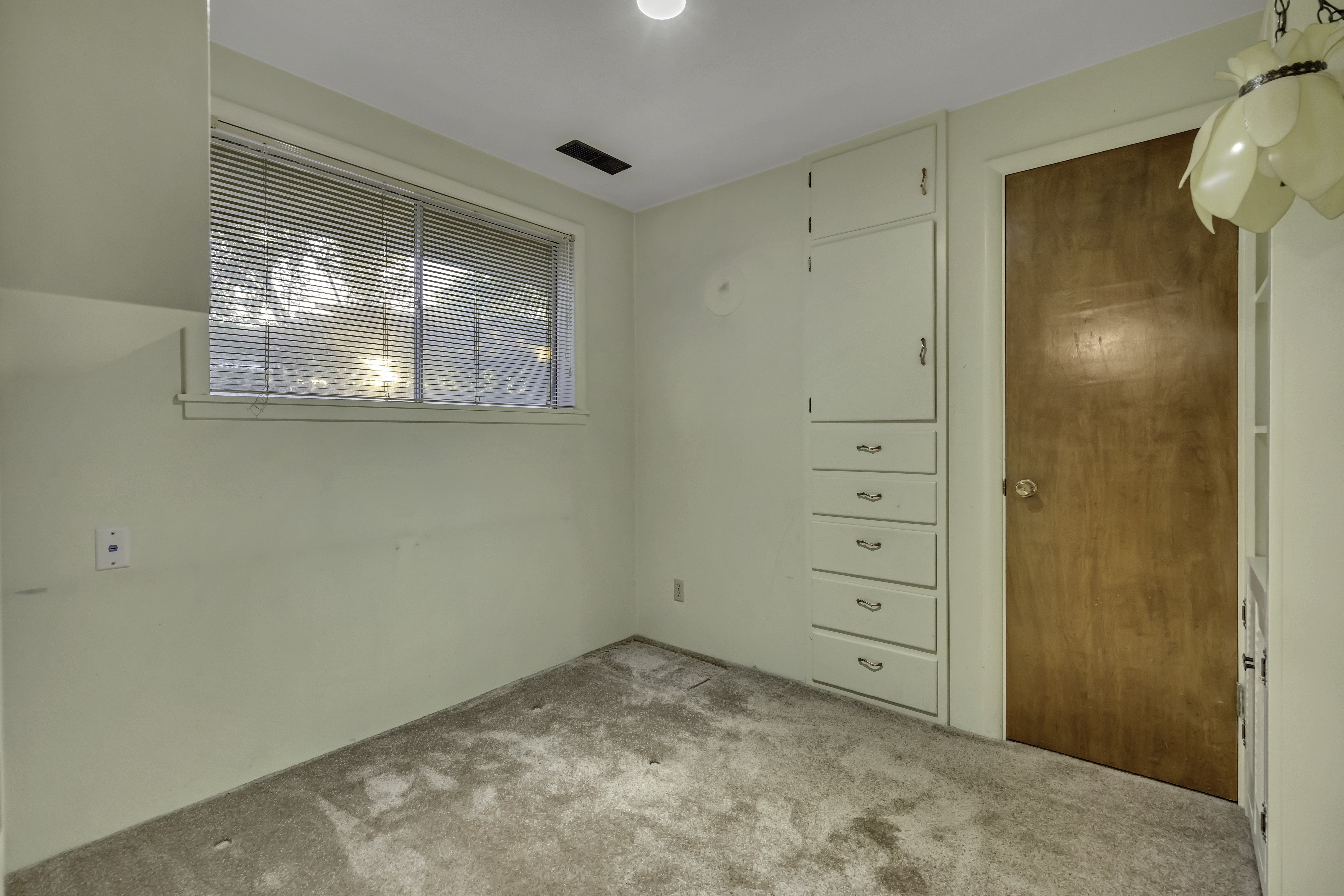 Room featuring a single window with blinds, light-toned carpeting, a built-in storage unit with drawers and an upper cabinet, and a natural wood-finish door