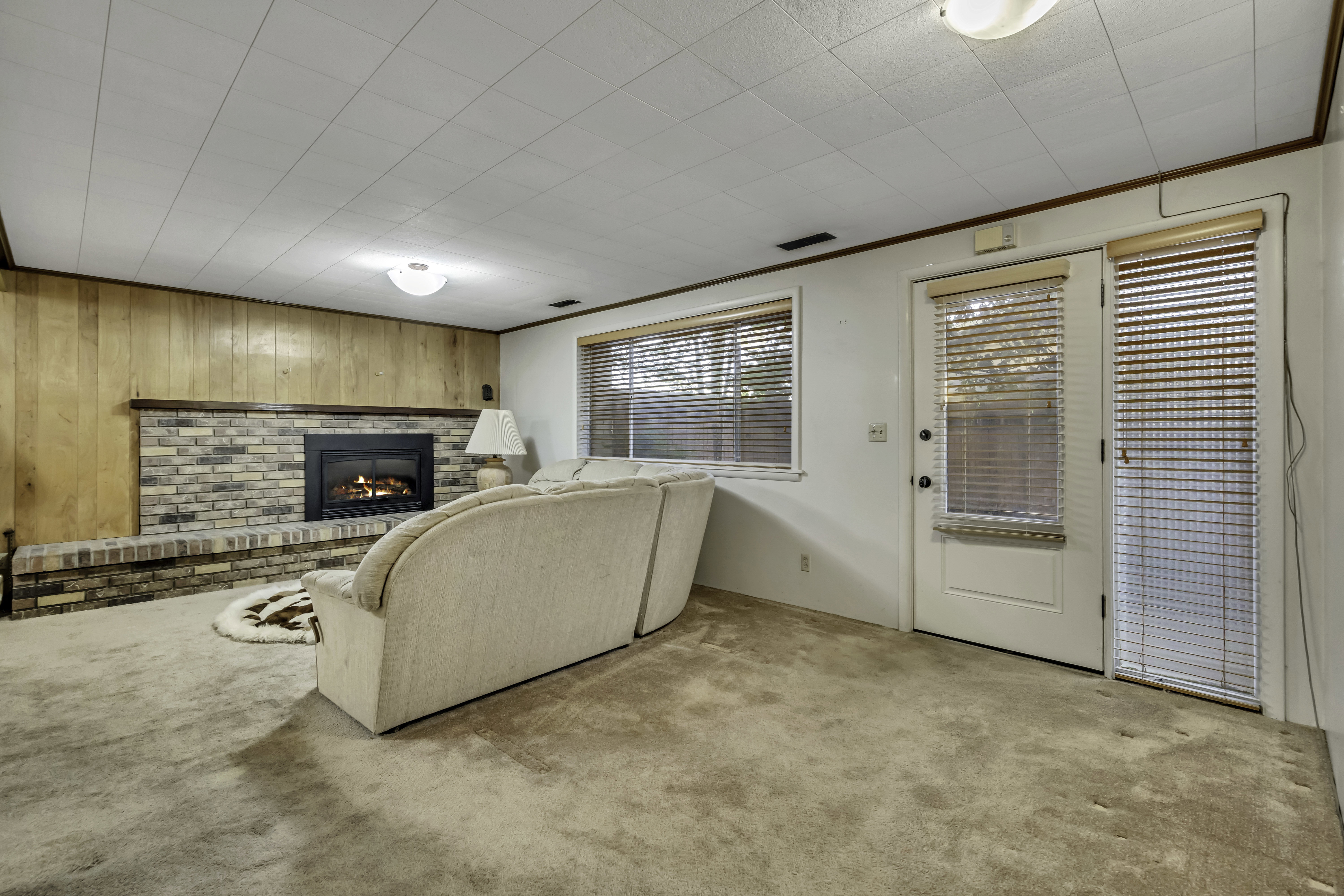 Spacious room featuring a brick fireplace with a wood mantle, wood paneling, and a large window with blinds
