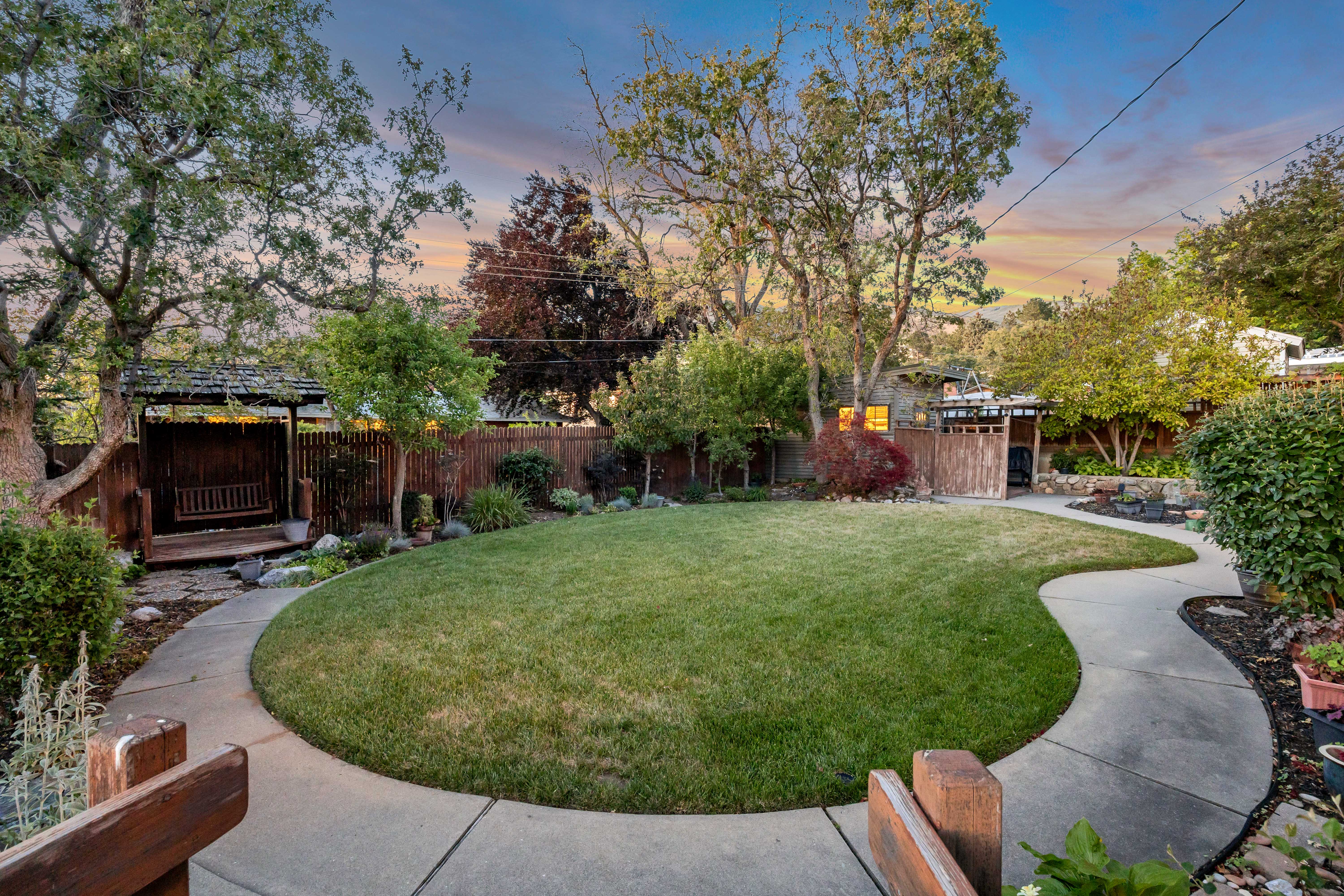 Expansive backyard featuring a large lawn area, meandering concrete pathway, mature trees, and a wooden gazebo structure