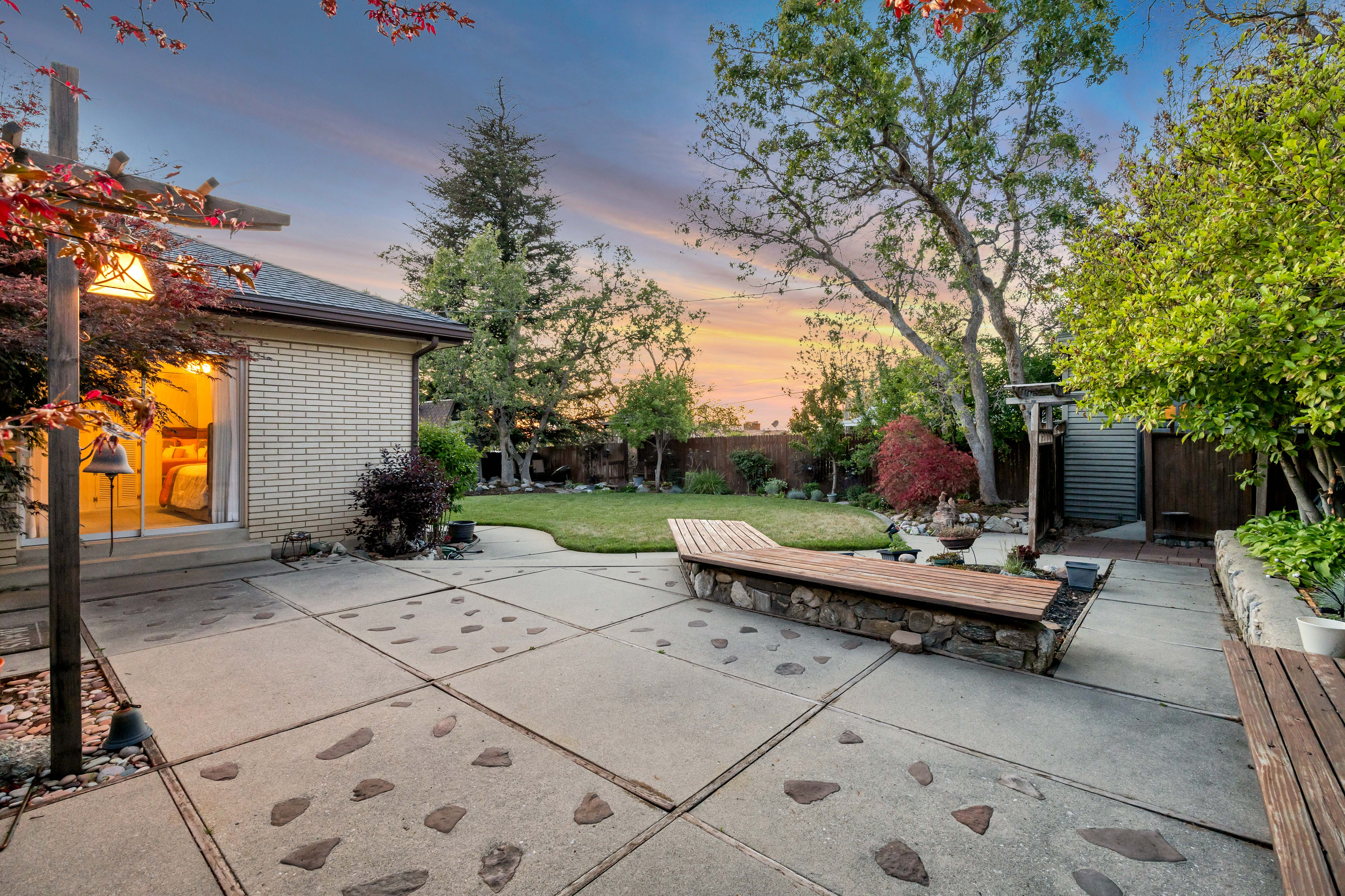 Expansive backyard featuring a large concrete patio with stone inlays, a built-in stone and wood bench, mature trees, and a manicured lawn