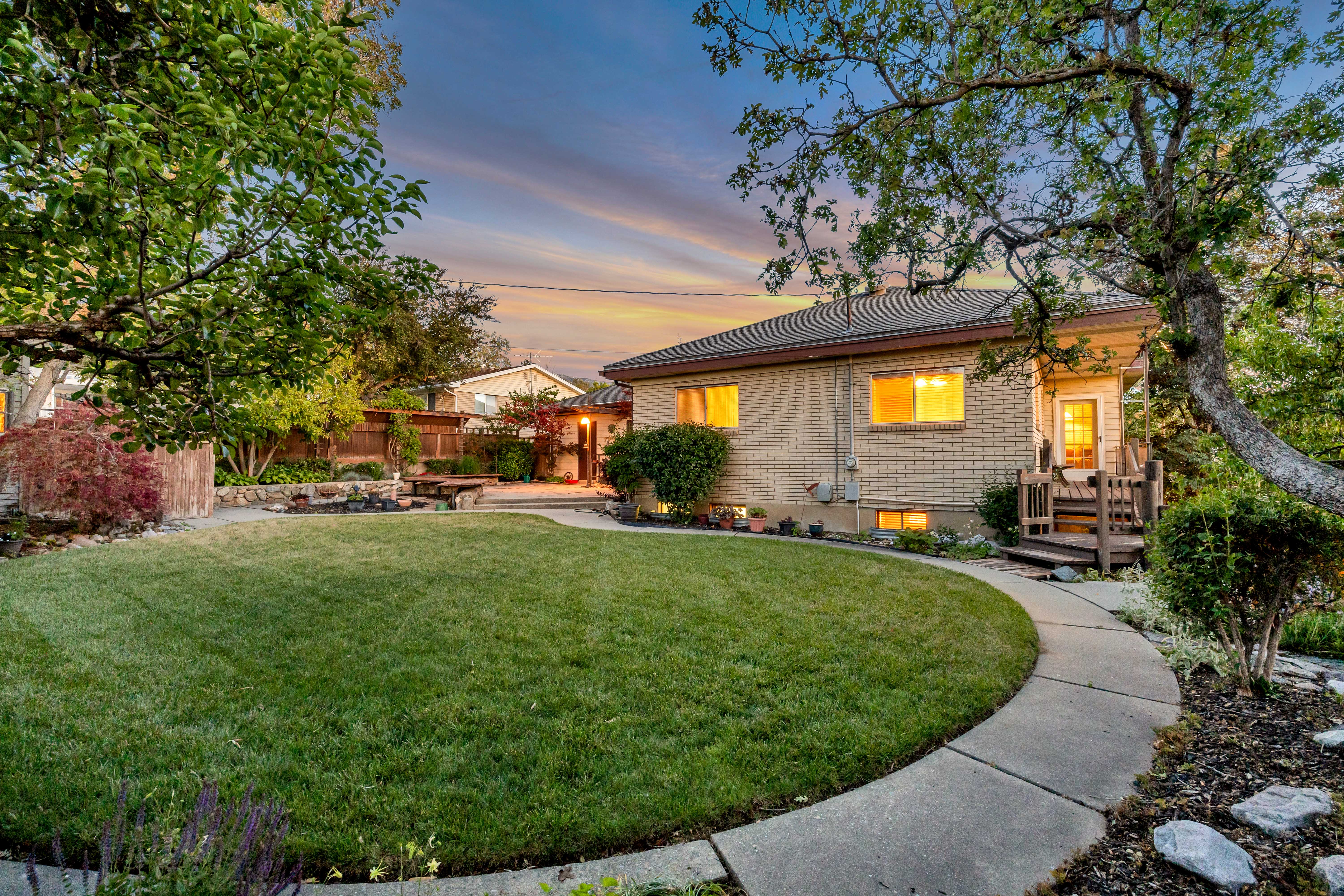 Expansive backyard featuring a manicured lawn, established trees, a curved concrete pathway, and a brick exterior dwelling with a wooden deck