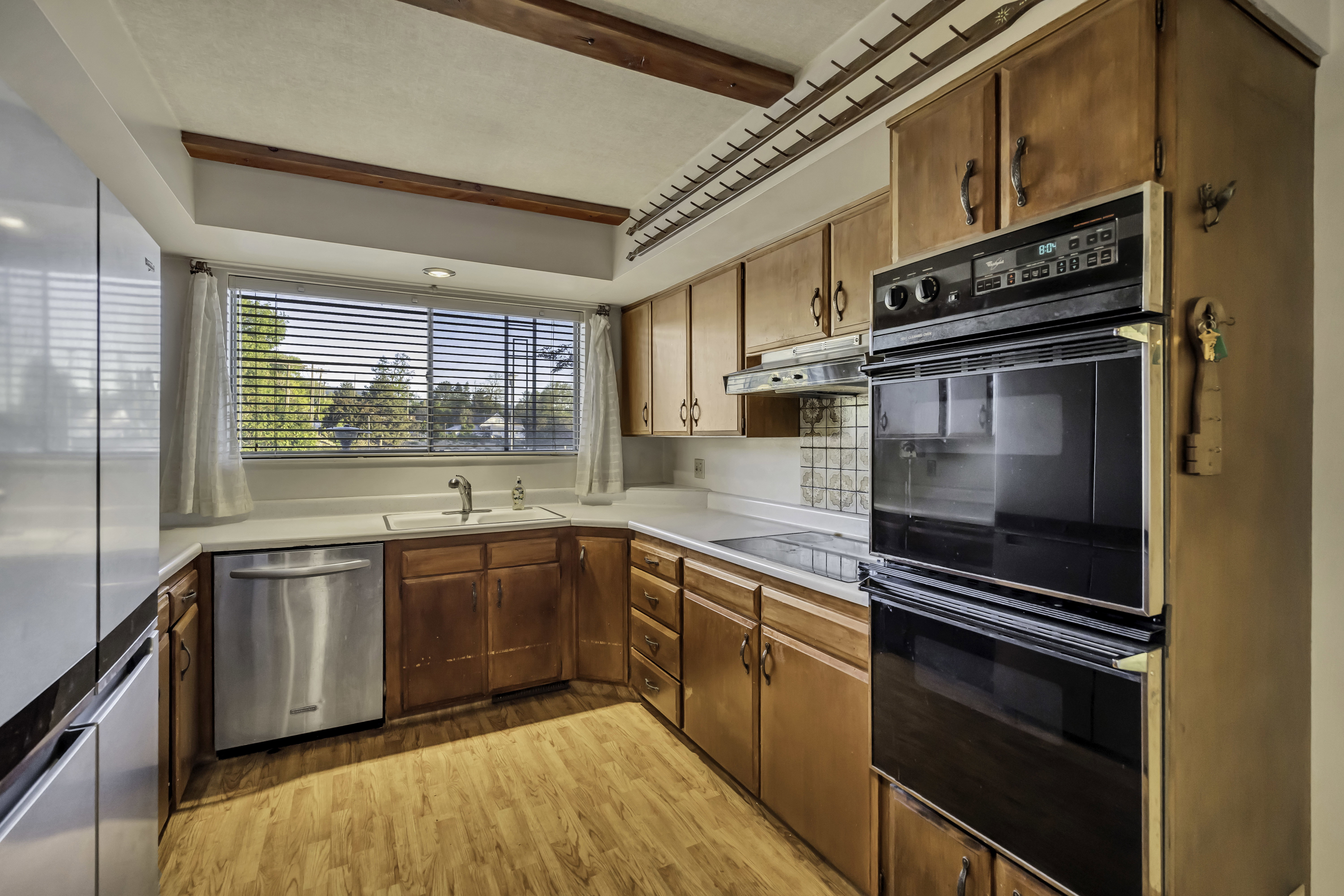 Kitchen featuring wood-finish flooring, wood cabinetry, light-toned countertops, a stainless steel dishwasher, and a double wall oven