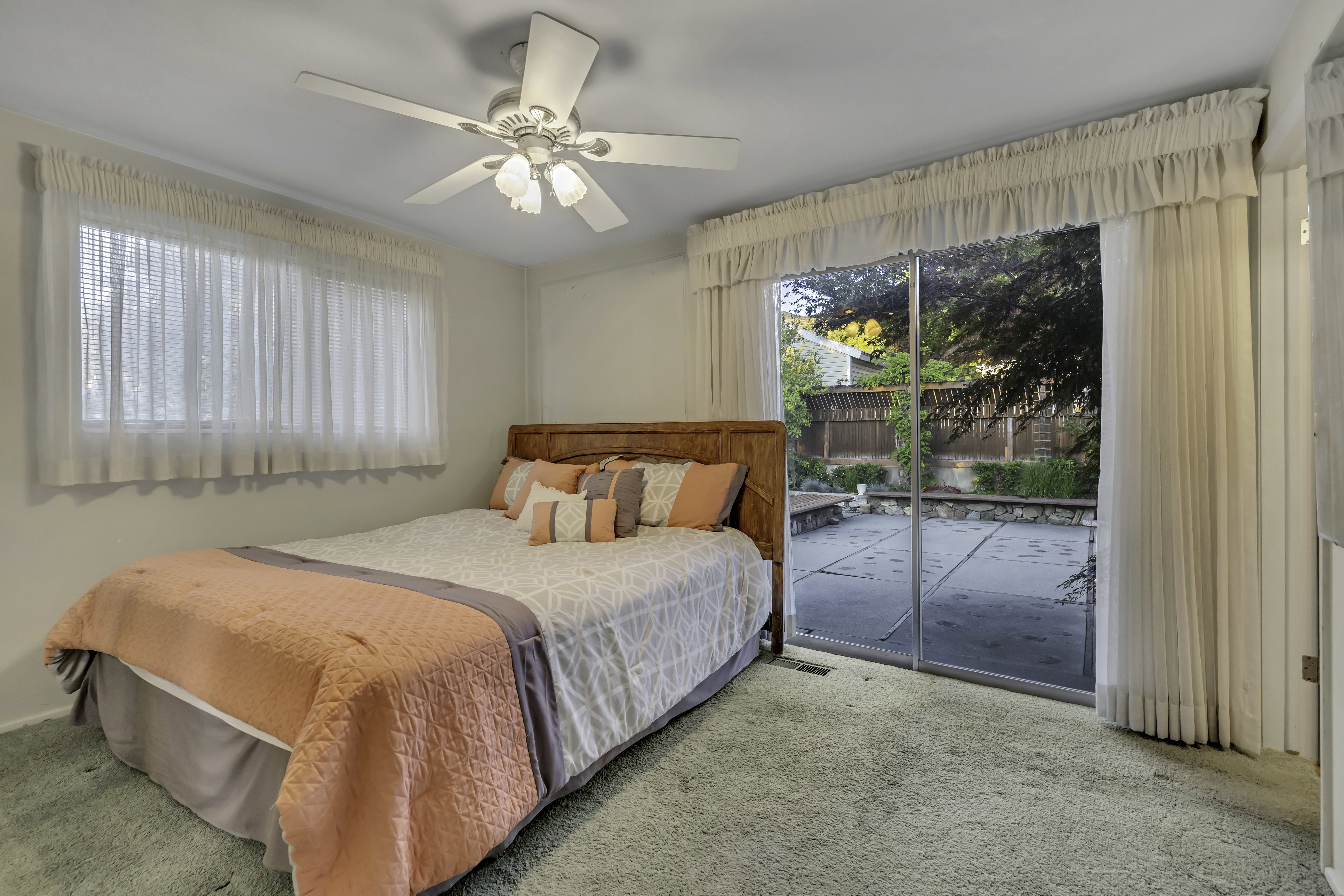Room featuring a ceiling fan with integrated lighting, large window with blinds and sheer curtains, and sliding glass doors providing outdoor access to a patio area