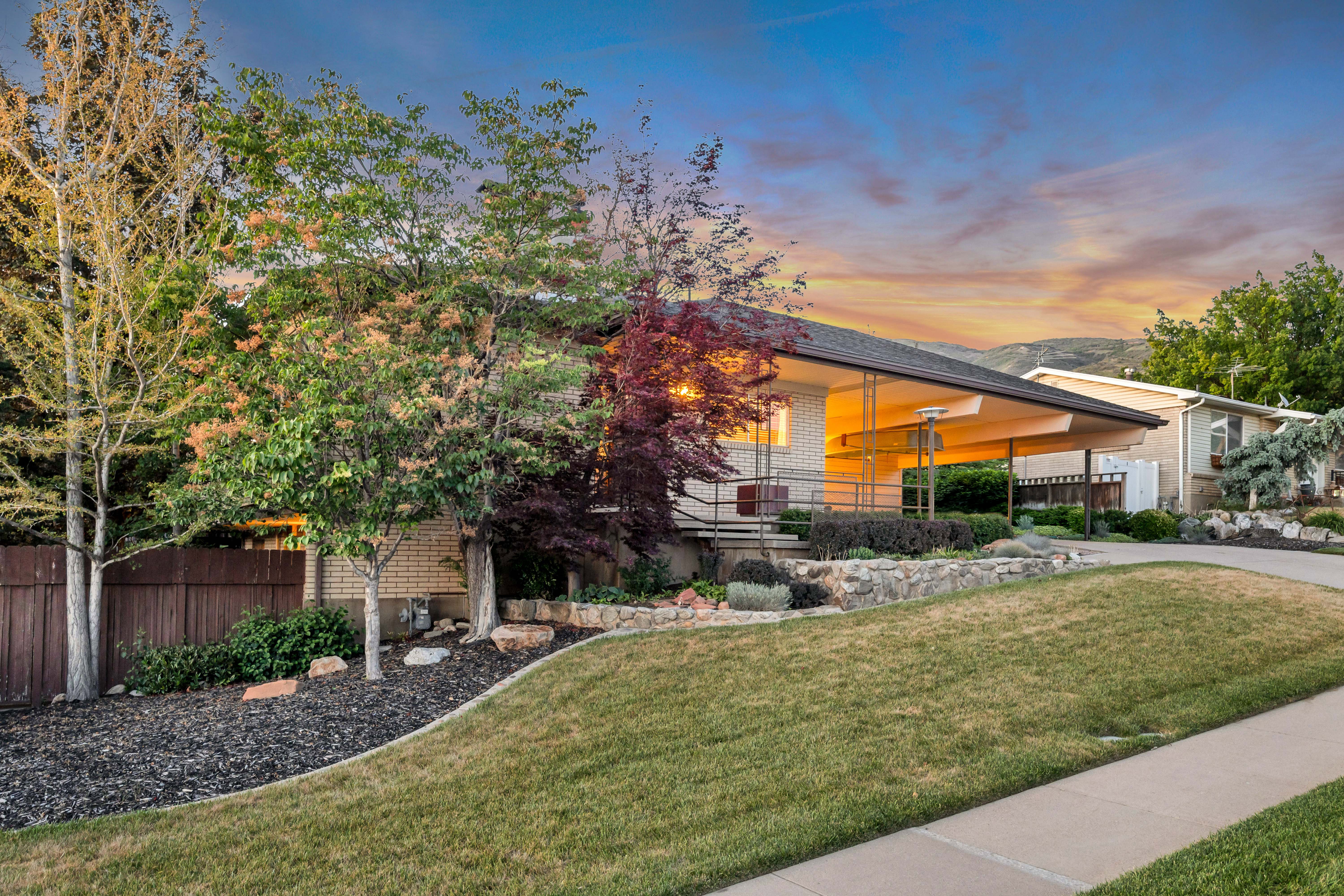 Brick facade residence with a covered carport, sloped terrain, mature trees, and a concrete driveway
