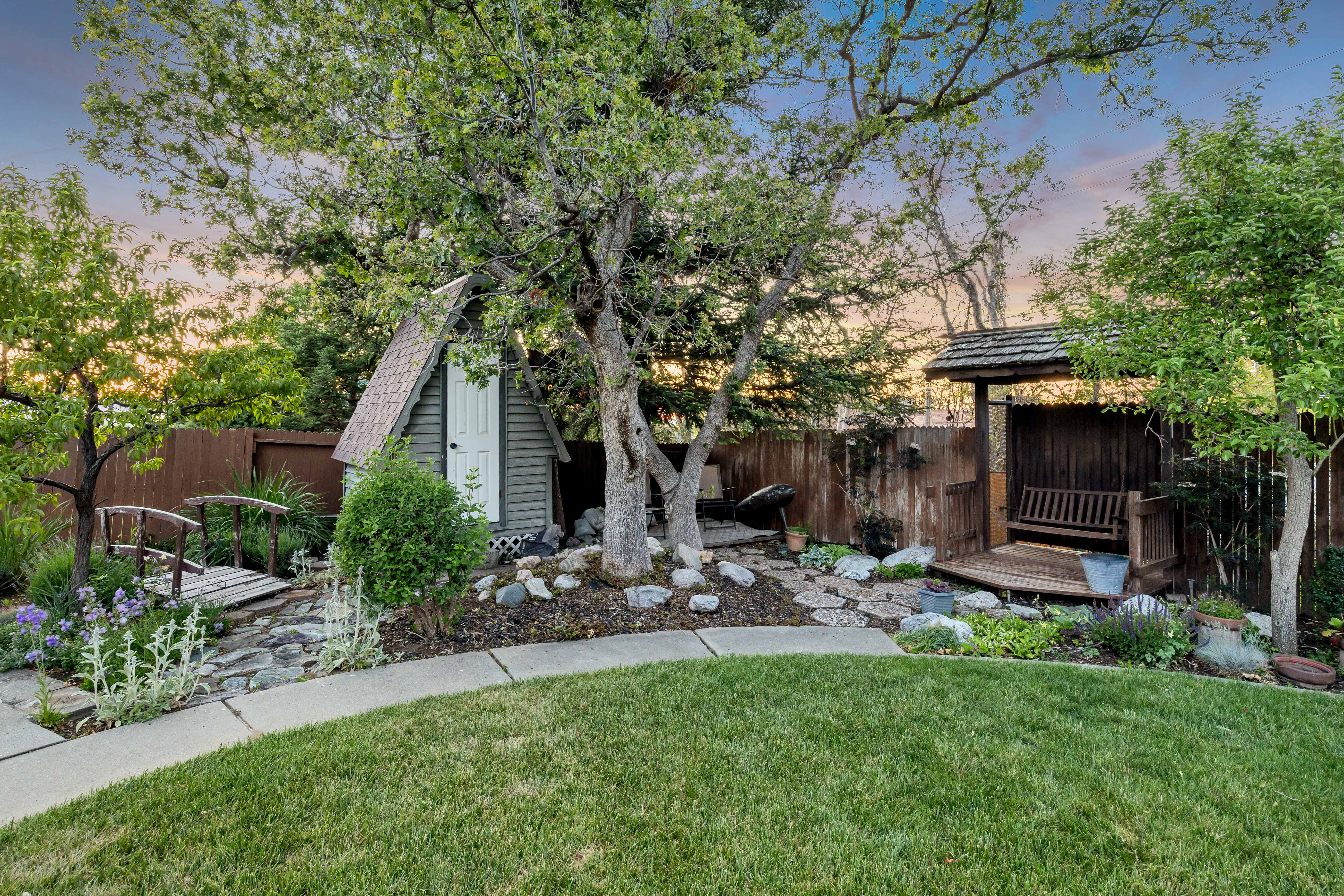 Manicured lawn with a curved concrete pathway leading to a landscaped garden featuring mature trees, a decorative bridge, and a rustic storage shed
