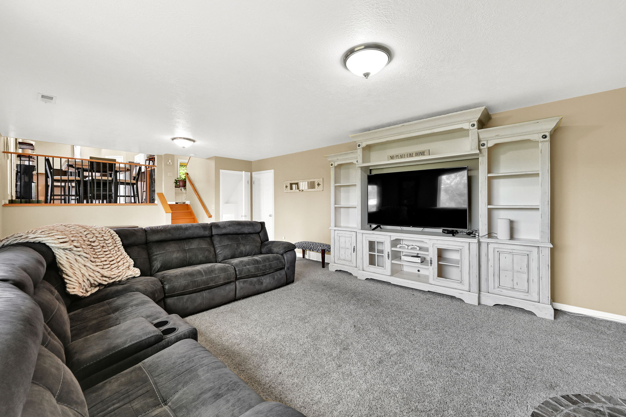 Carpeted living area featuring a built-in media console with shelving and cabinet storage, ceiling-mounted lighting, and a partial view of a staircase with wood treads and a railing