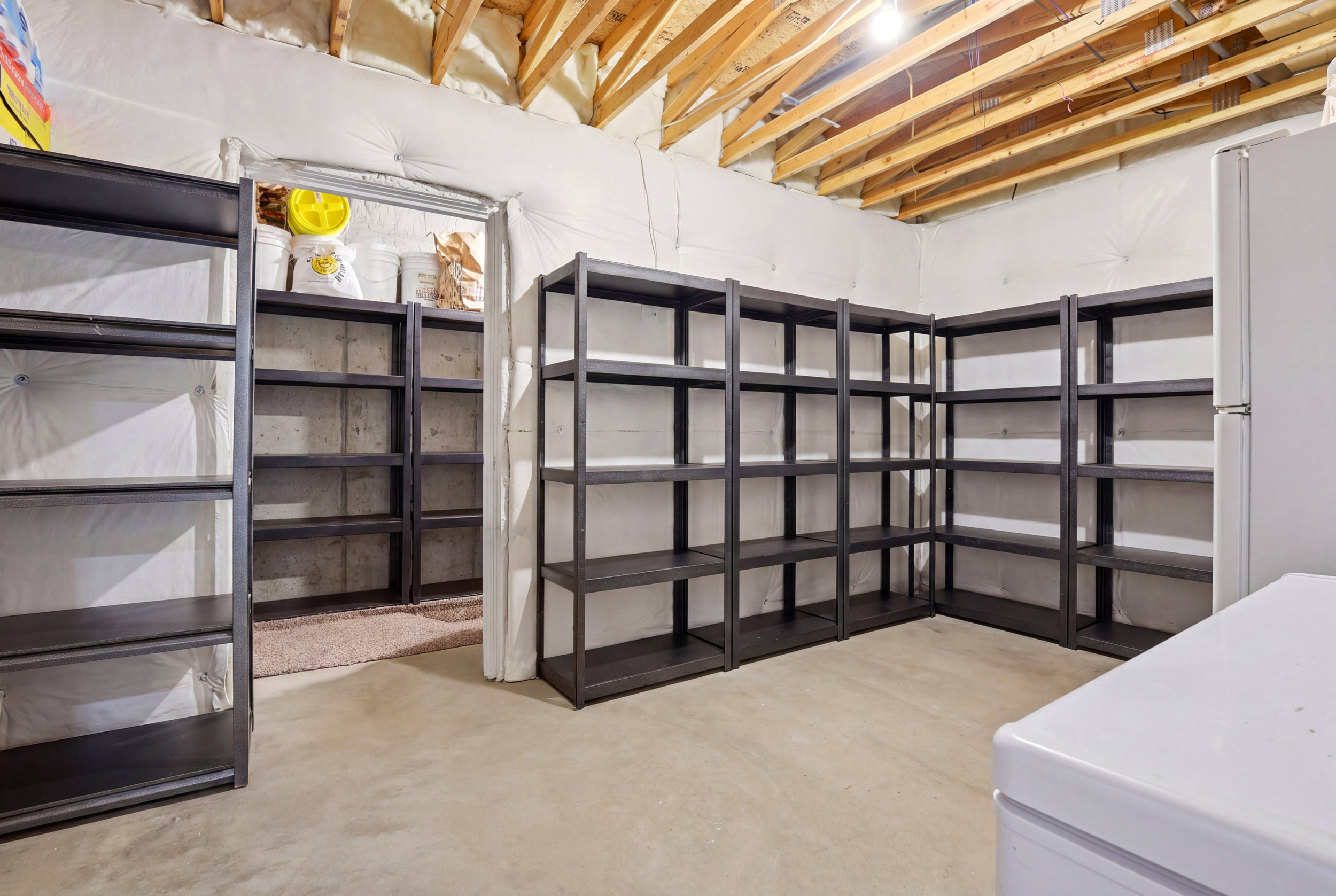 Utility room featuring extensive built-in shelving, exposed ceiling joists, concrete flooring, and an additional storage area