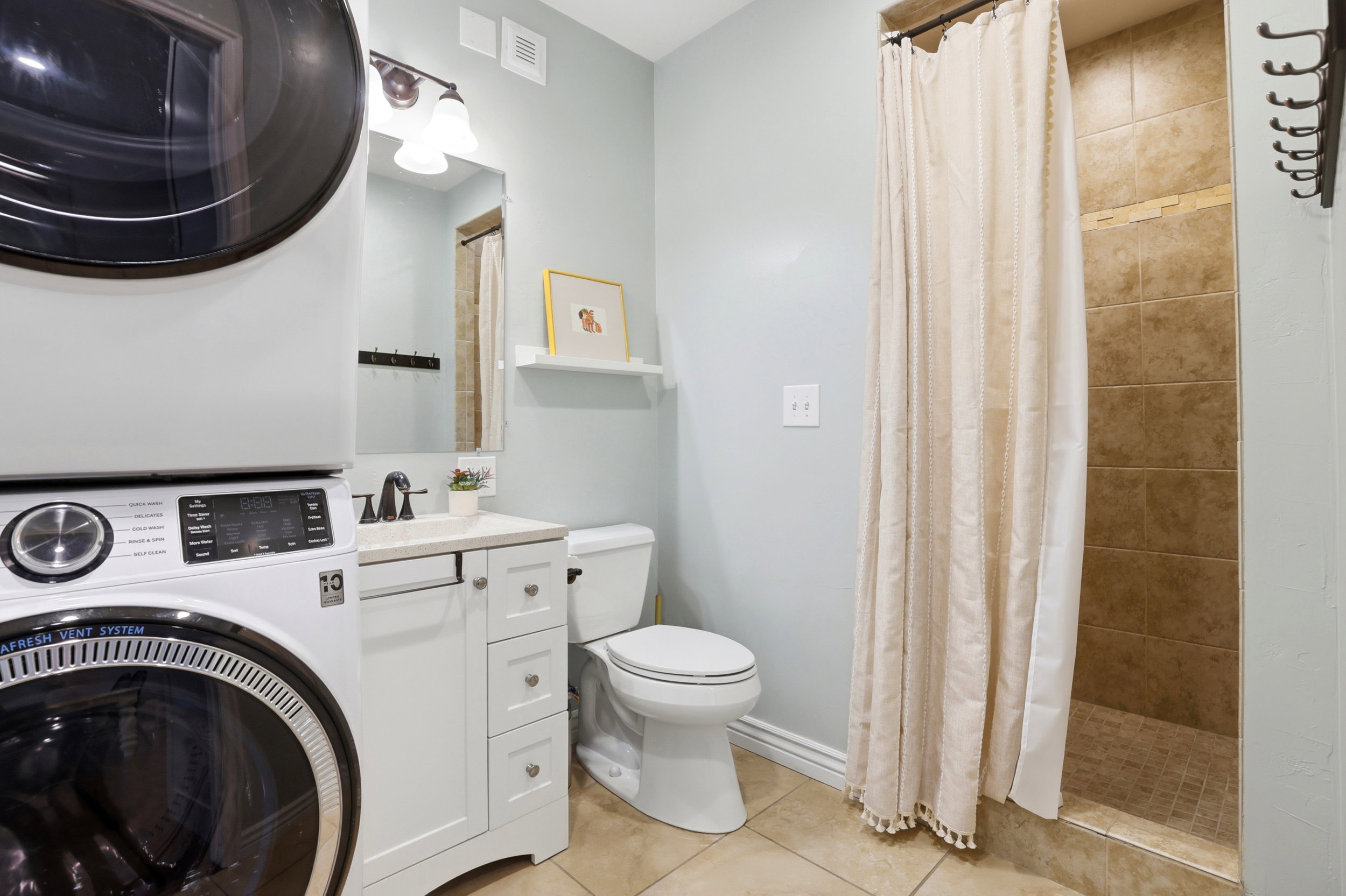 Bathroom featuring a stacked washer and dryer, a white vanity with a countertop, a toilet, a wall-mounted mirror, and a tiled shower stall