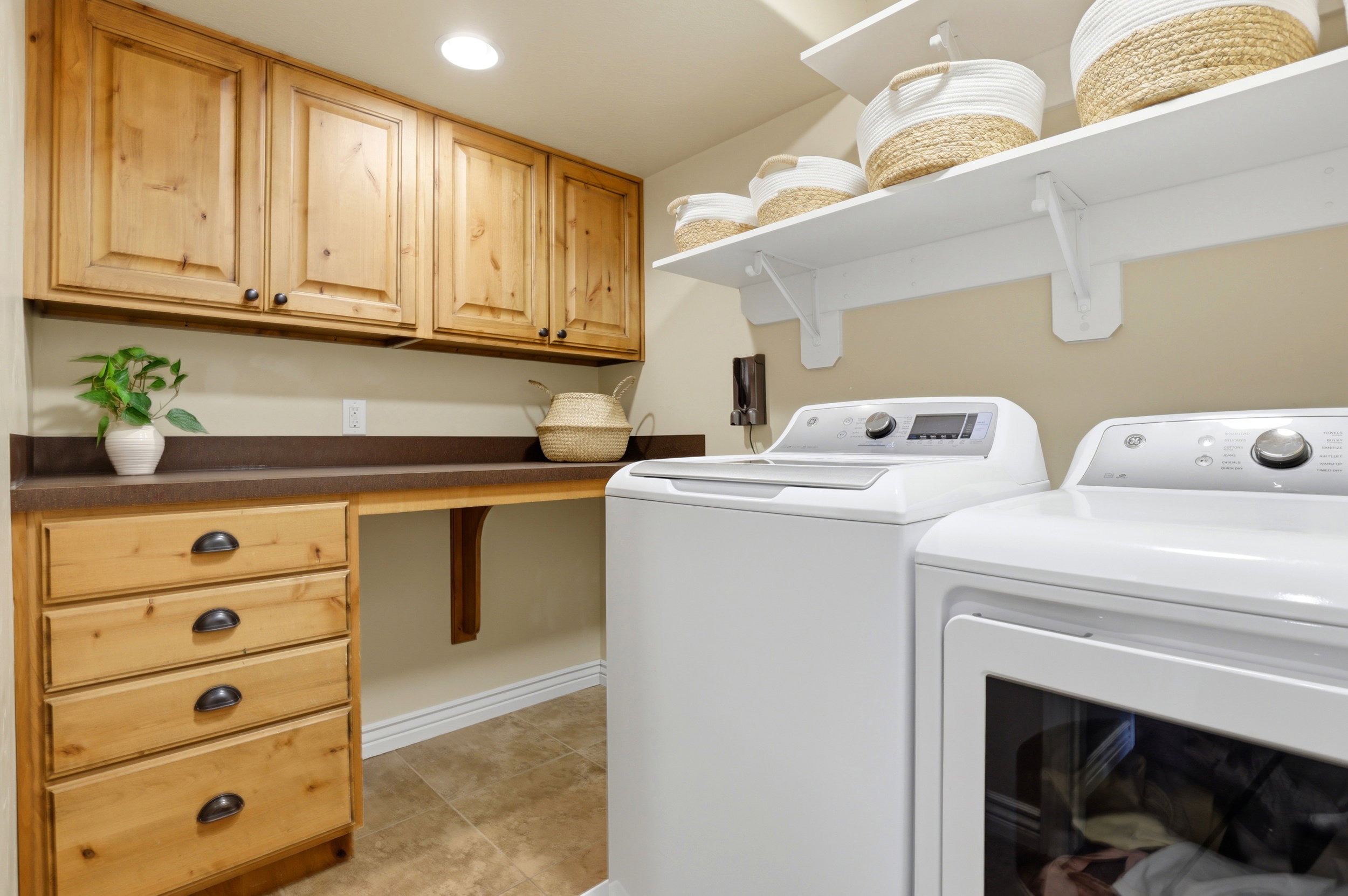 Laundry area featuring natural wood cabinetry, integrated countertop space, upper storage shelving, and tile flooring