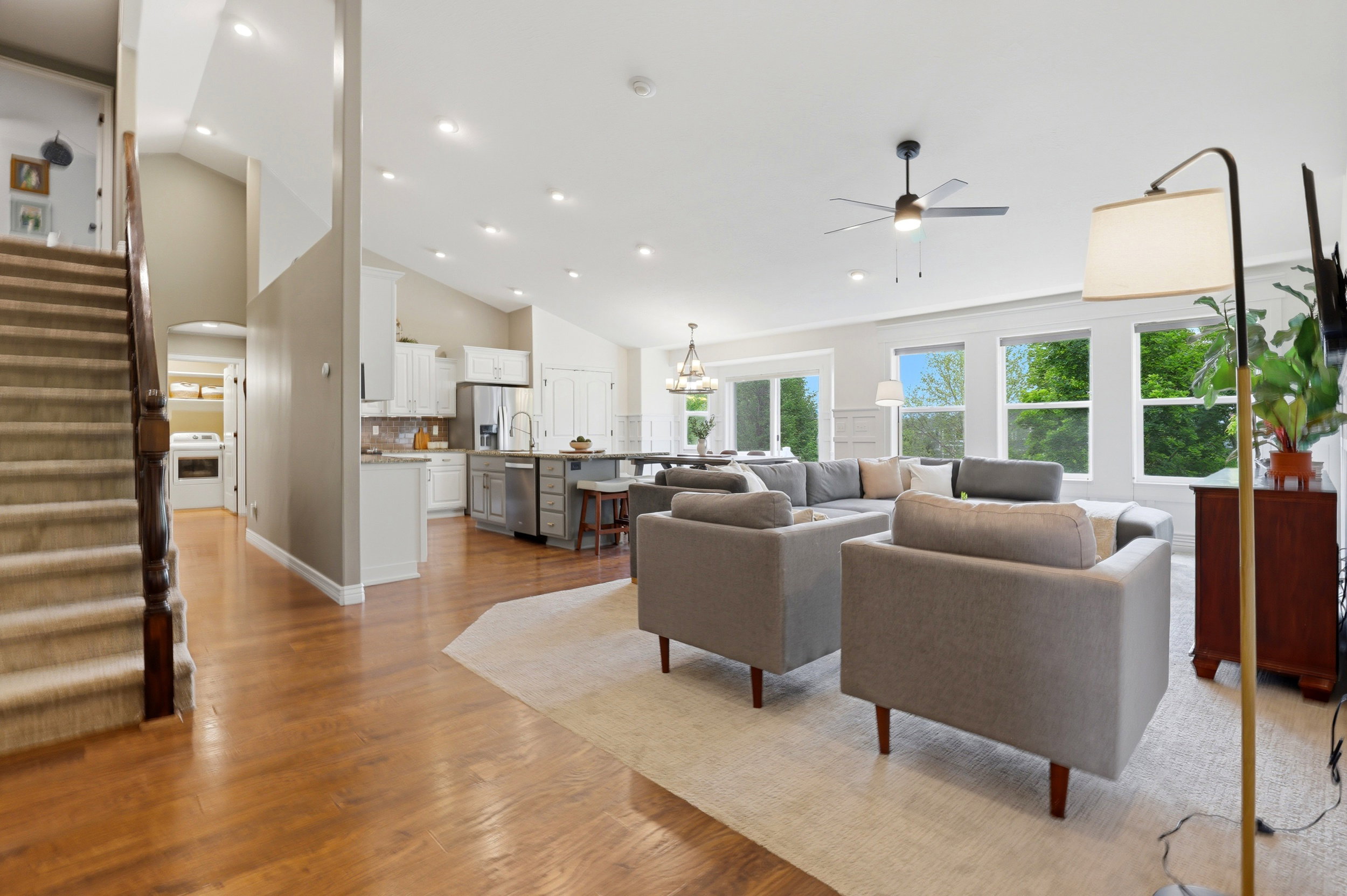 Spacious open-concept interior featuring vaulted ceilings, wood-finish flooring, white cabinetry, a kitchen island with seating, and multiple large windows