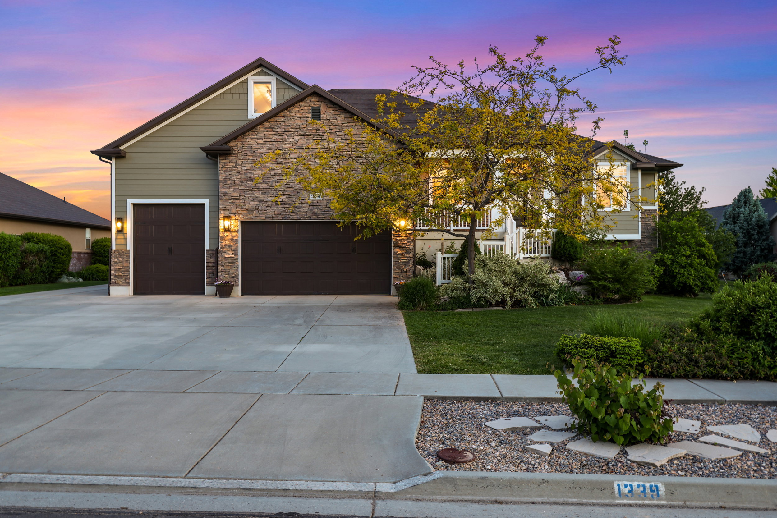 Stone and siding exterior featuring a multi-car garage, a concrete driveway, and a landscaped front yard with mature trees and shrubbery