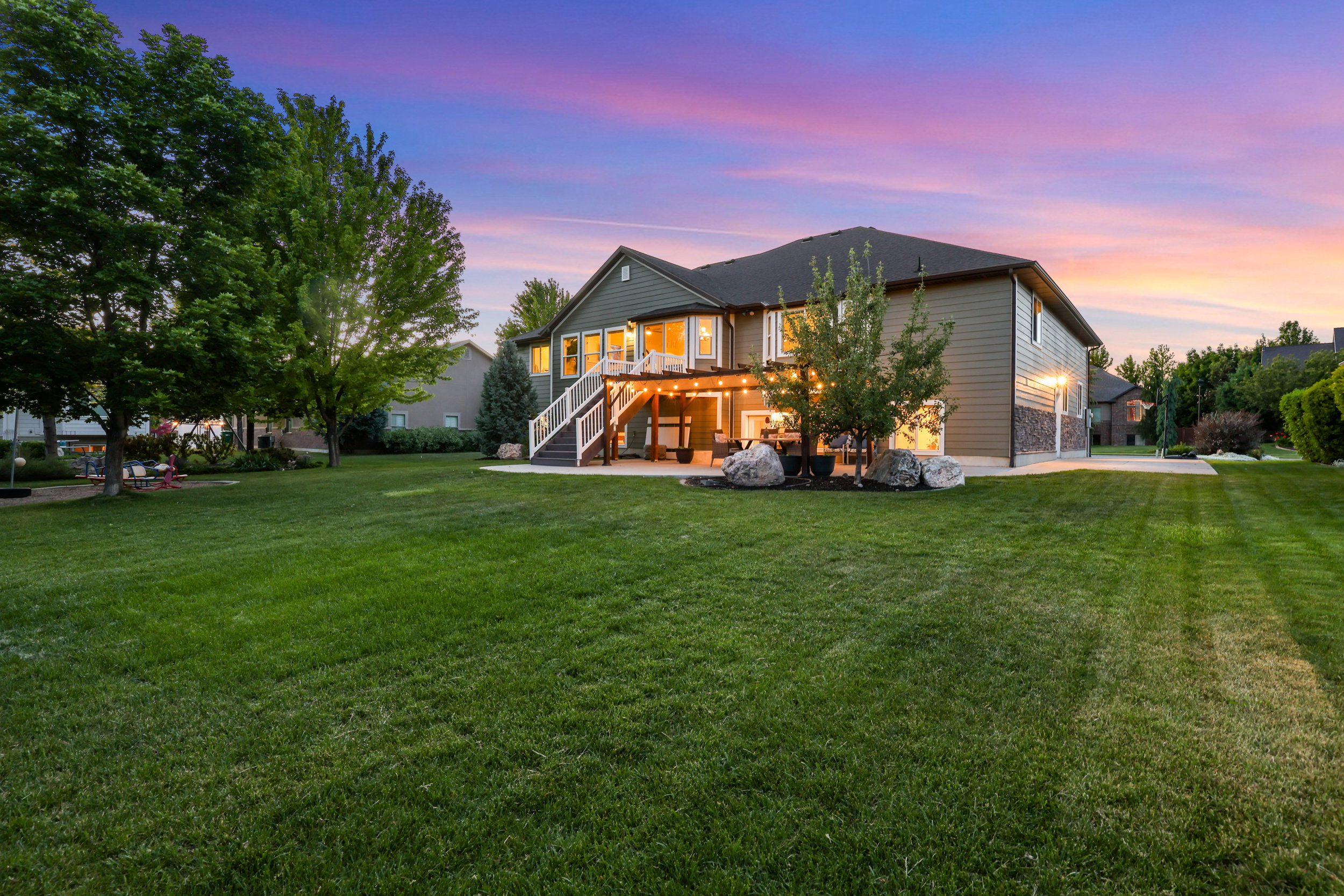 Expansive green lawn with mature trees, a two-story home featuring a second-story deck, a covered patio, and stone accents on the lower facade