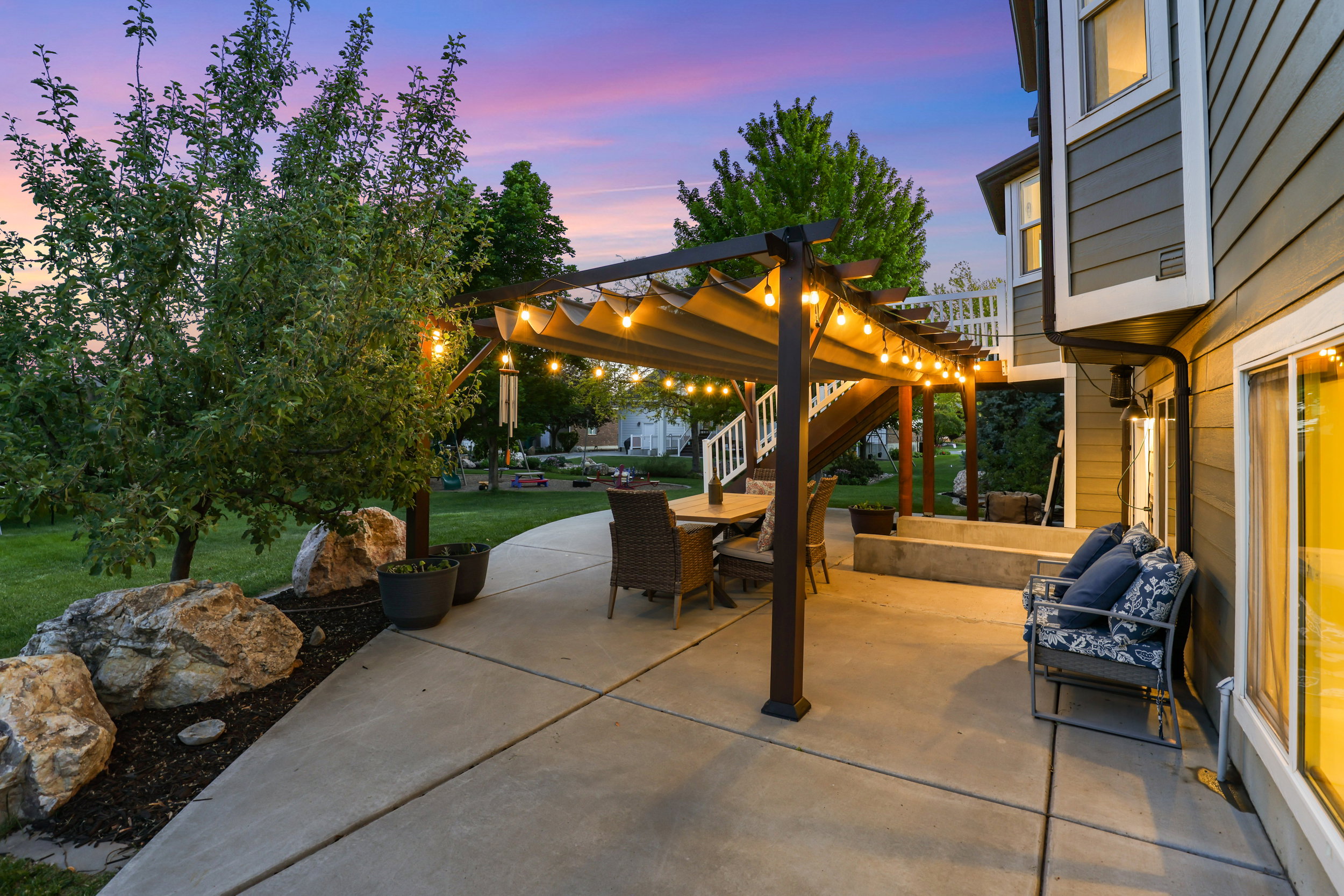 Concrete patio with a pergola featuring string lighting, a manicured lawn, and mature trees