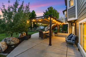 Concrete patio with a pergola featuring string lighting, a manicured lawn, and mature trees