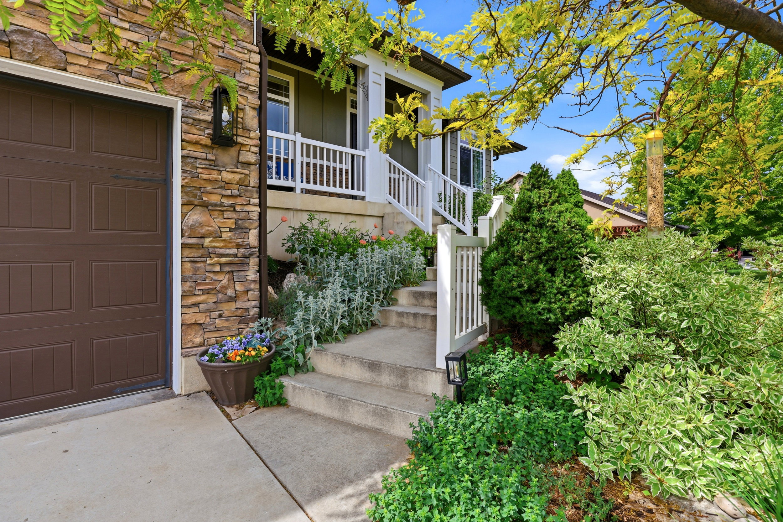 Front exterior featuring a brown paneled garage door, stacked stone siding, concrete steps, white railings, and lush landscaping