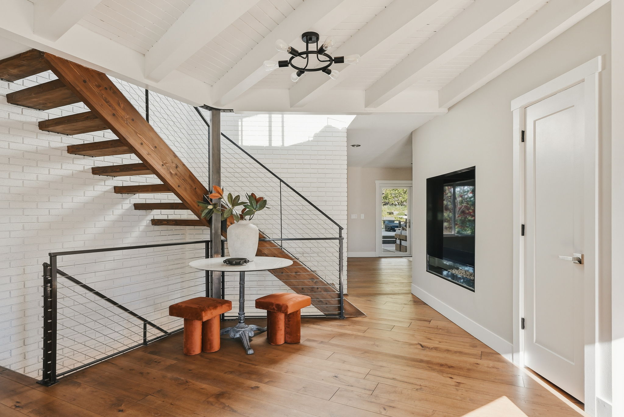 Open-riser staircase with wood treads and metal cable railing, white painted brick accent wall, wood-finish flooring, recessed linear fireplace, and vaulted ceiling with exposed beams