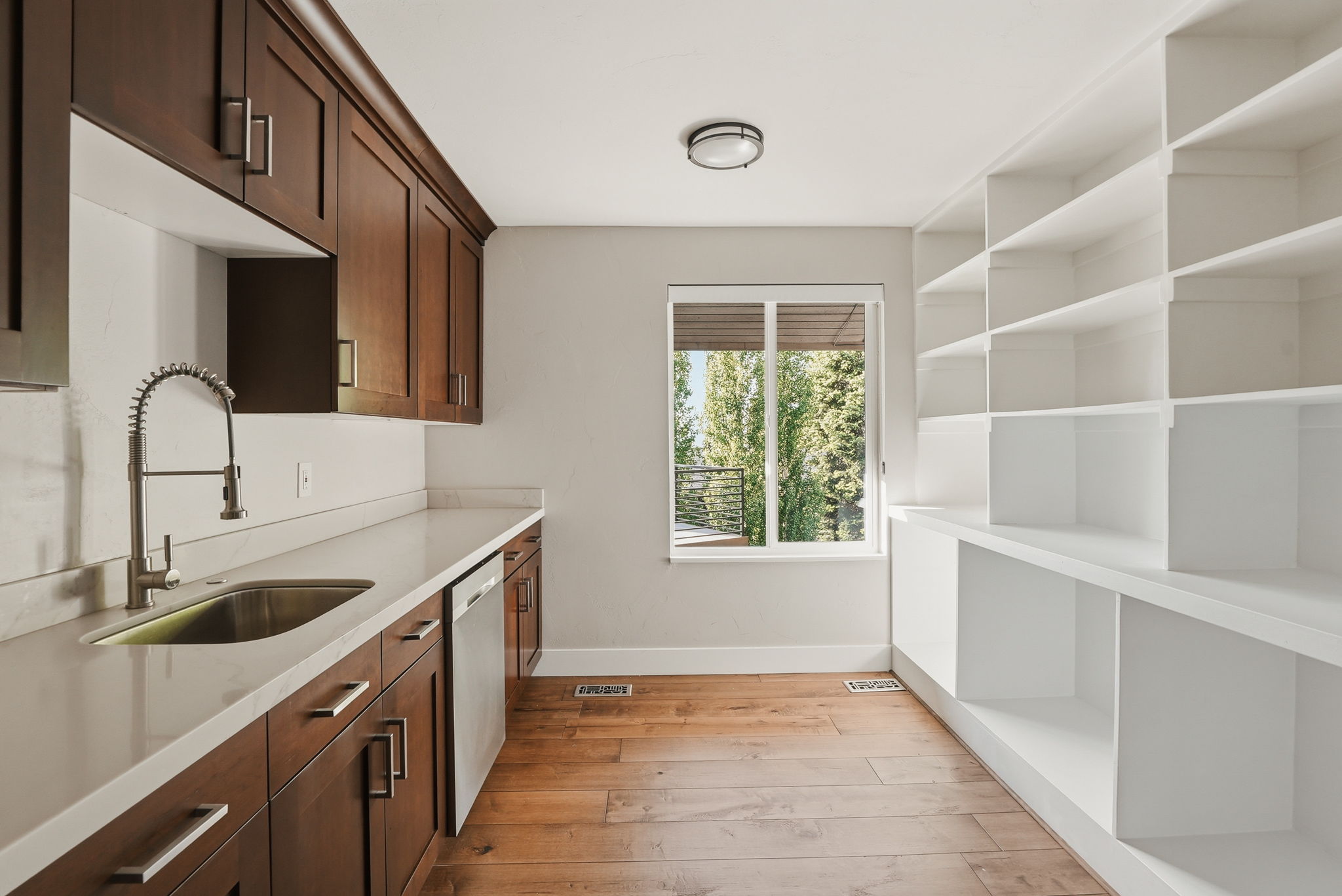 Spacious pantry featuring dark wood-finish cabinetry, light-toned countertops, a stainless steel sink with a gooseneck faucet, an integrated dishwasher, and extensive white built-in shelving