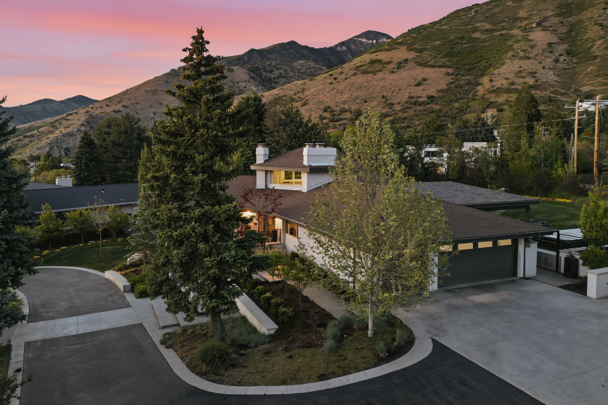 Modern residential architecture with a neutral exterior palette, mature landscaping, a dark asphalt driveway, a two-car garage, and a prominent mountain backdrop