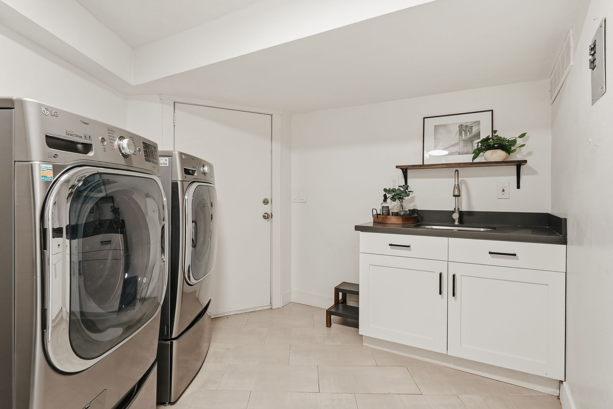 Laundry area with light tile flooring, white walls, built-in white cabinetry, a dark countertop with an undermount sink, and a wall-mounted wooden shelf