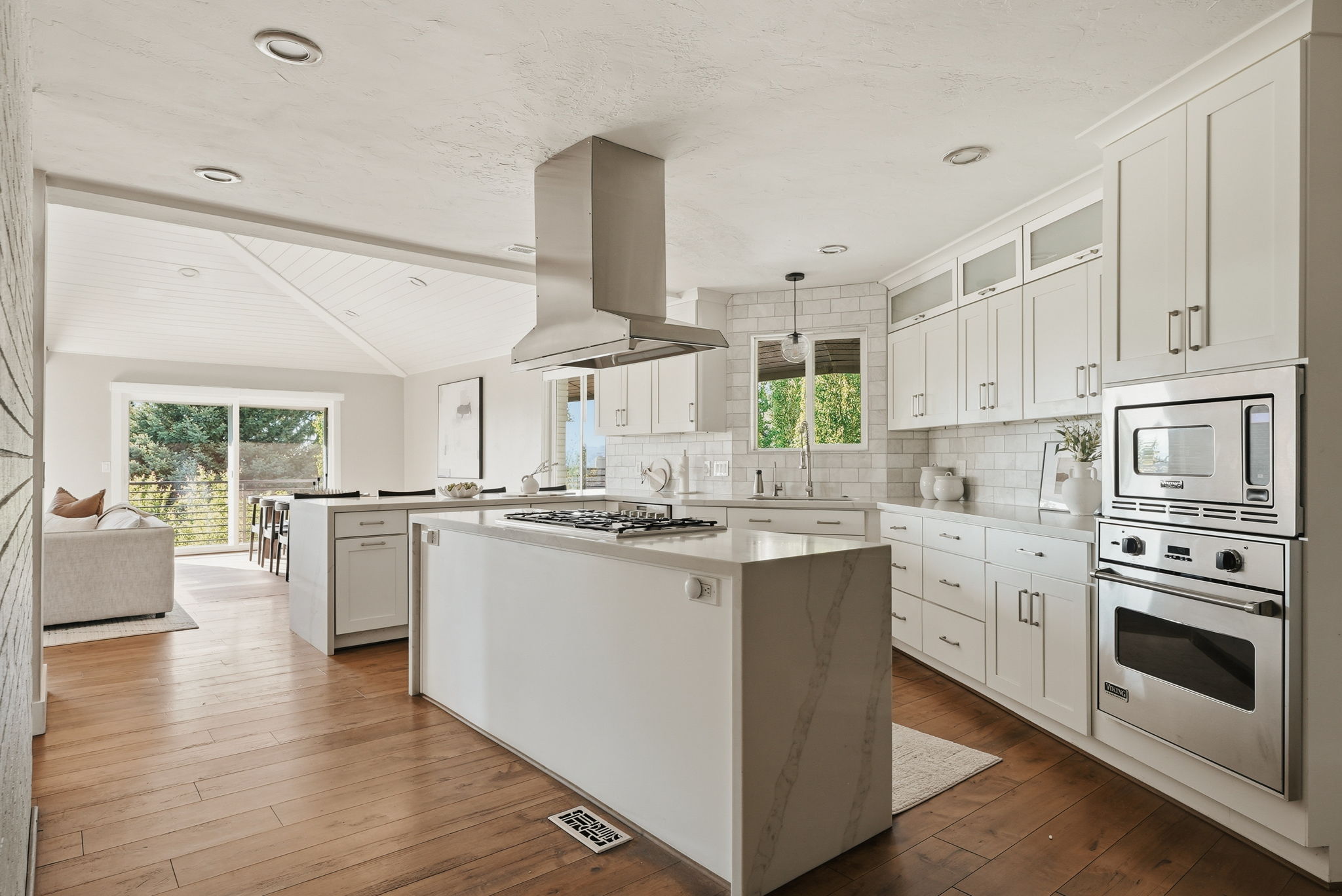Gourmet kitchen featuring a waterfall island, stainless steel range hood, built-in wall oven and microwave, white shaker cabinetry, and wood-finish flooring