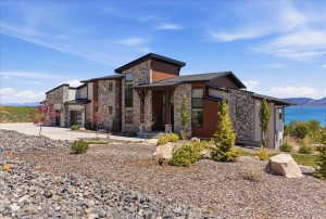 Contemporary residence featuring stone and wood siding, multiple garages, a covered front entry, and a prominent water backdrop