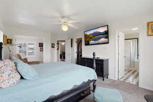 Carpeted bedroom featuring a ceiling fan with integrated lighting, an open doorway to an adjoining room with window blinds, and an open doorway to a bathroom with checkered tile flooring and a shower enclosure