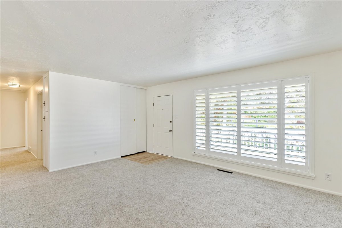 Bright living area featuring a wide window with plantation shutters, light-toned carpeting, and a white entry door