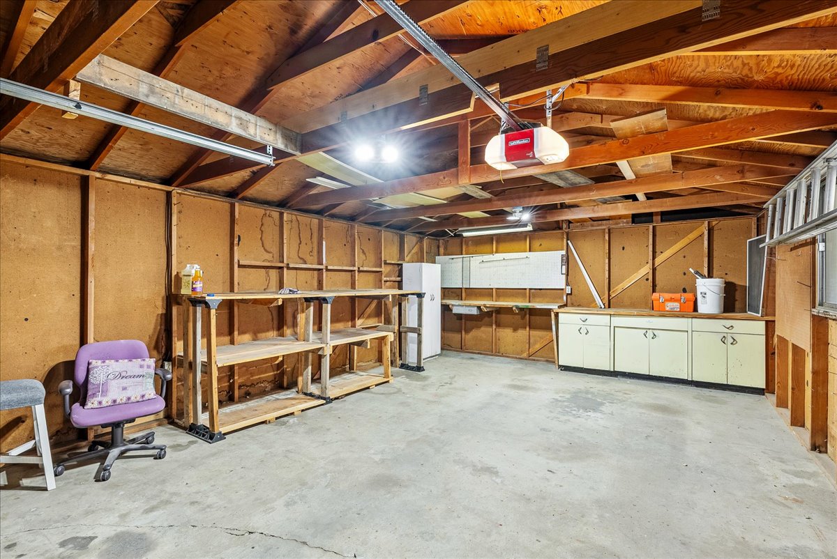 Spacious garage featuring exposed wood beam ceiling, concrete flooring, built-in shelving, wall-mounted cabinets, and overhead lighting