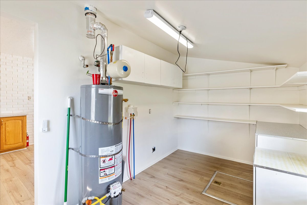 Dedicated utility area featuring a water heater, white built-in shelving, white overhead cabinetry, light wood-finish flooring, and a white ceiling with flush-mount lighting