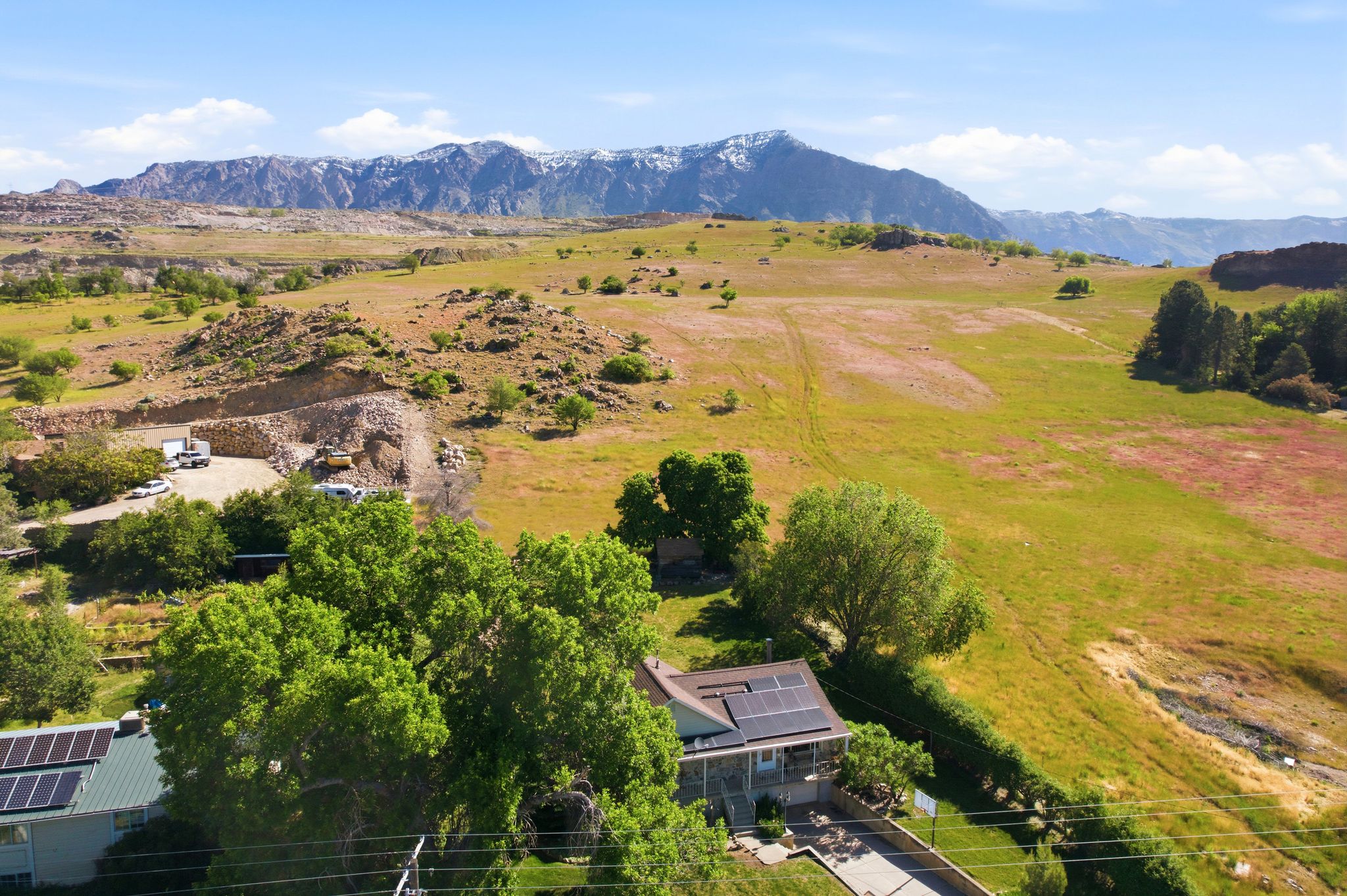 Expansive property featuring a residence with solar panels, mature trees, and a concrete driveway, set against a backdrop of rolling hills and distant snow-capped mountains
